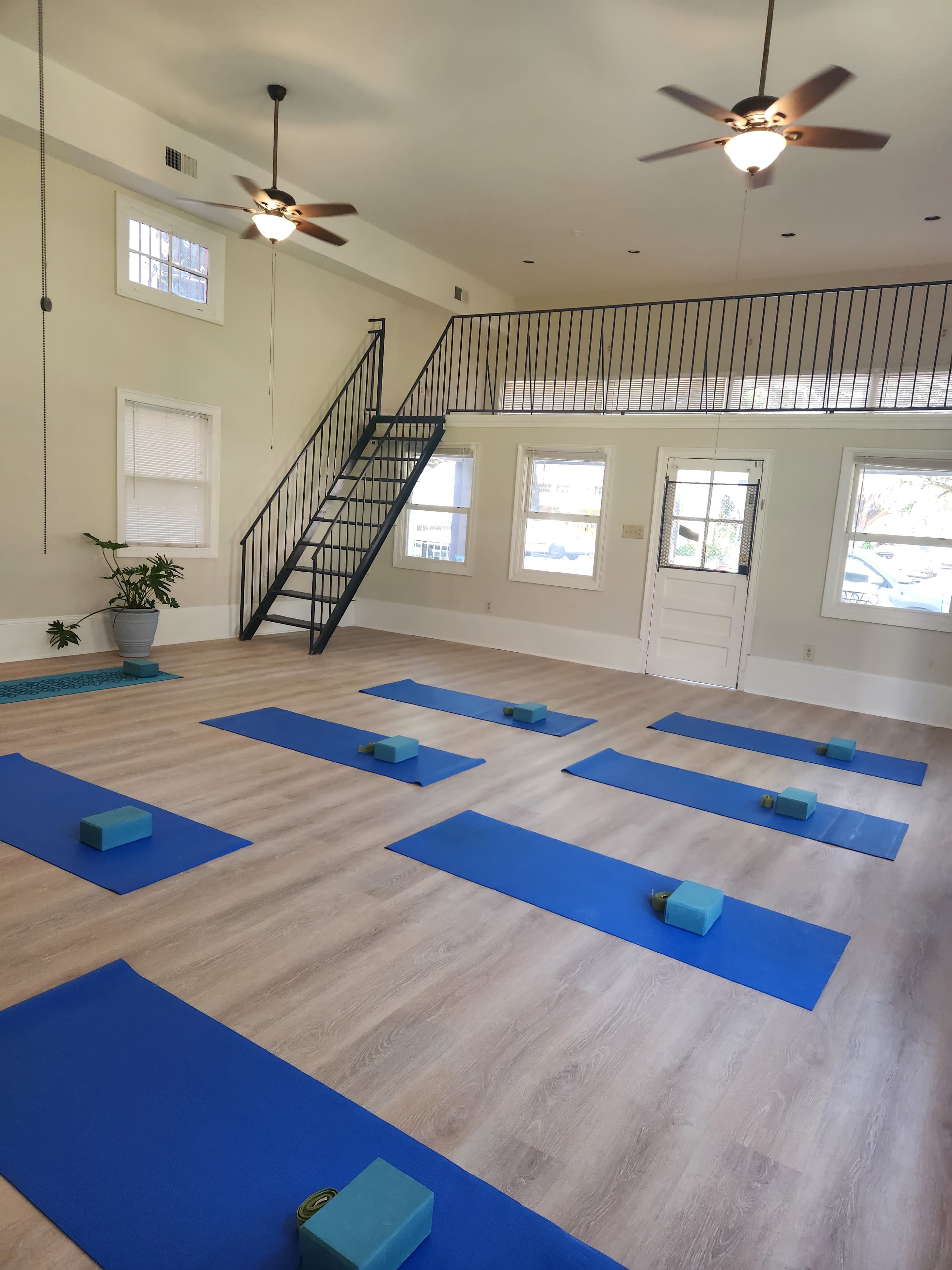 A spacious yoga studio with multiple blue mats and blocks arranged on the wooden floor, illuminated by ceiling fans and natural light from the windows.