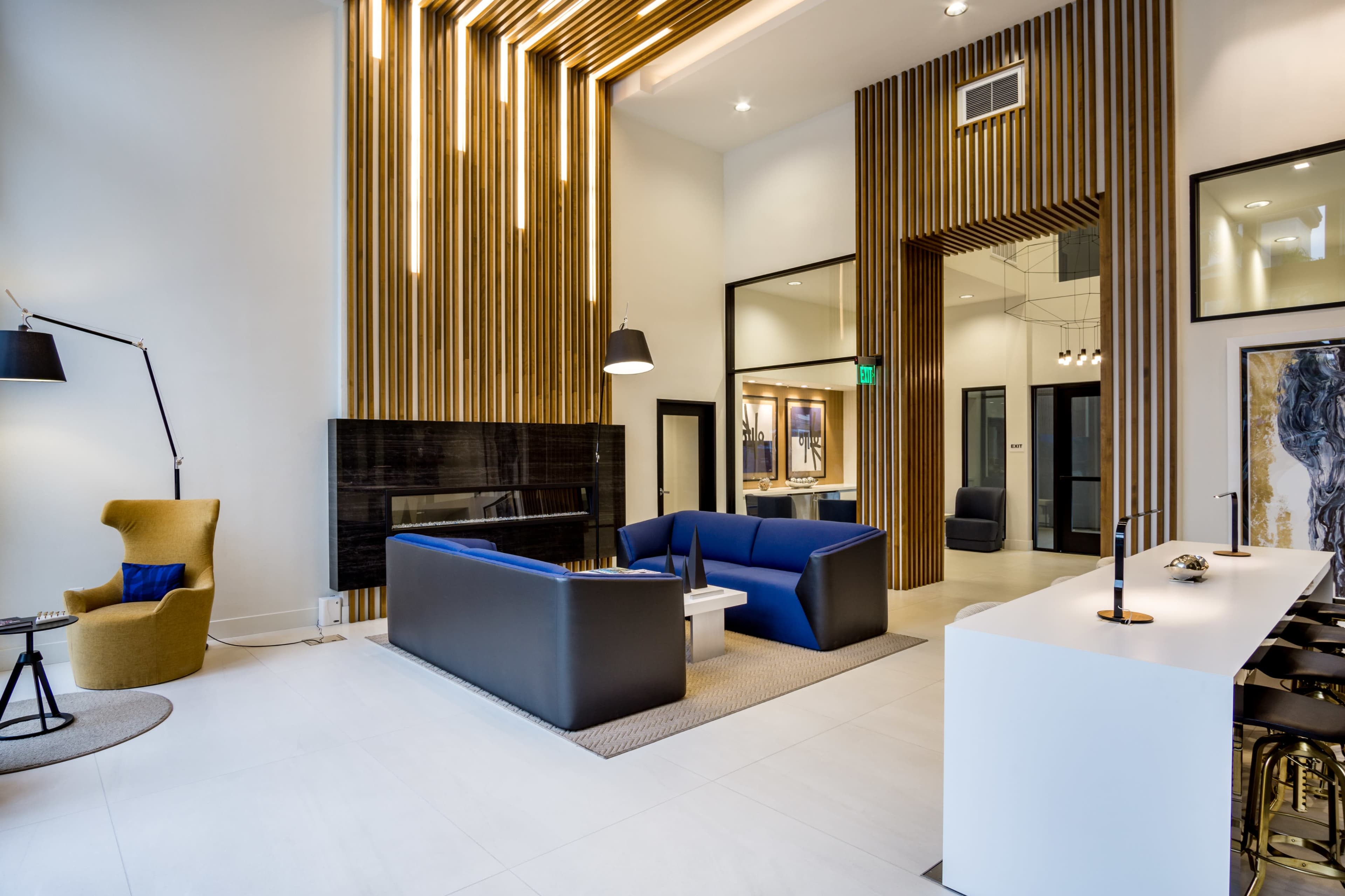 A modern lobby with wooden wall accents, a seating area including a blue couch and yellow chair, a minimalist bar, and large glass doors.