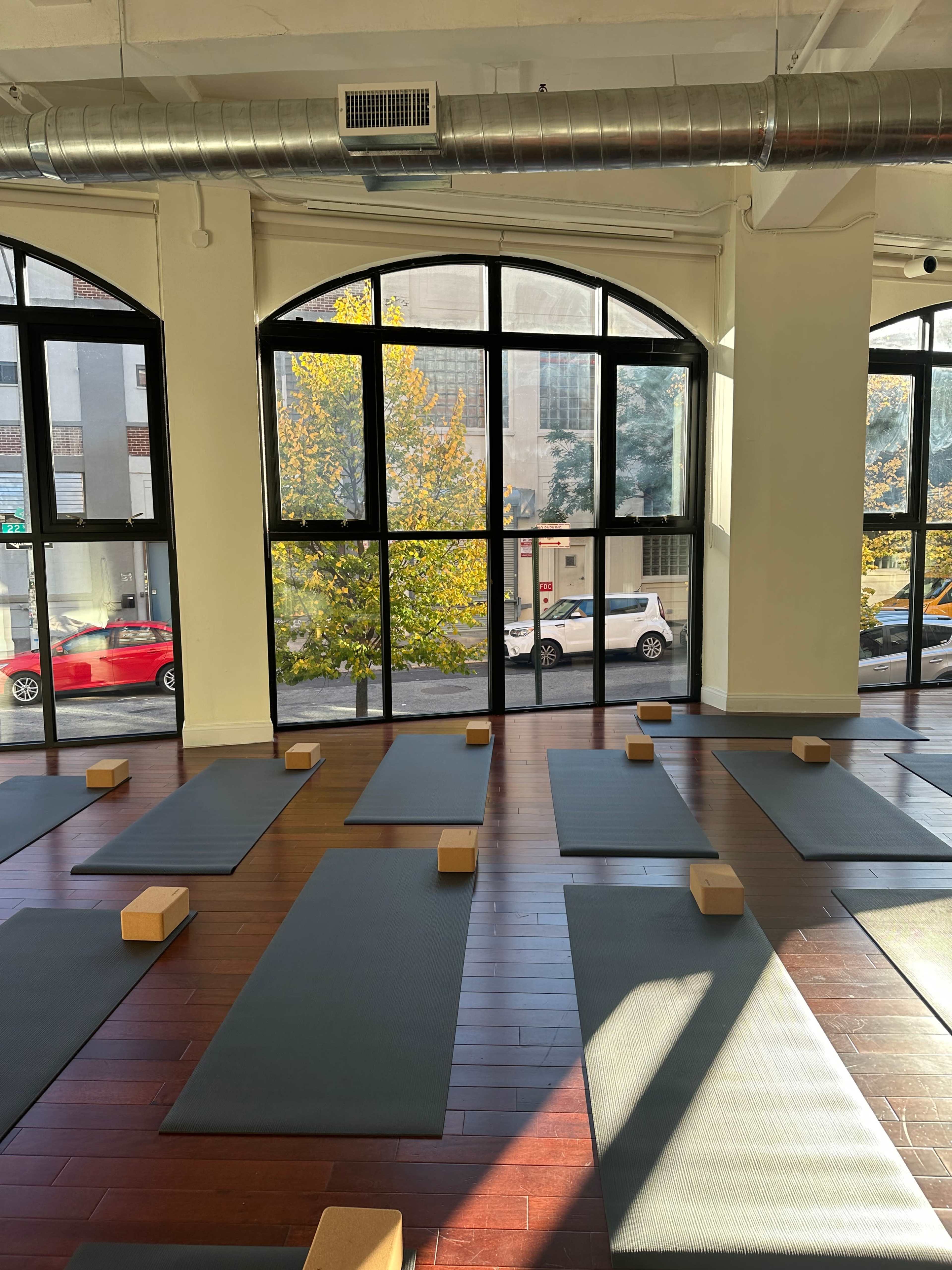 A yoga studio features several mats arranged on a wooden floor, with blocks placed on each mat and large windows revealing a cityscape and autumn trees outside.