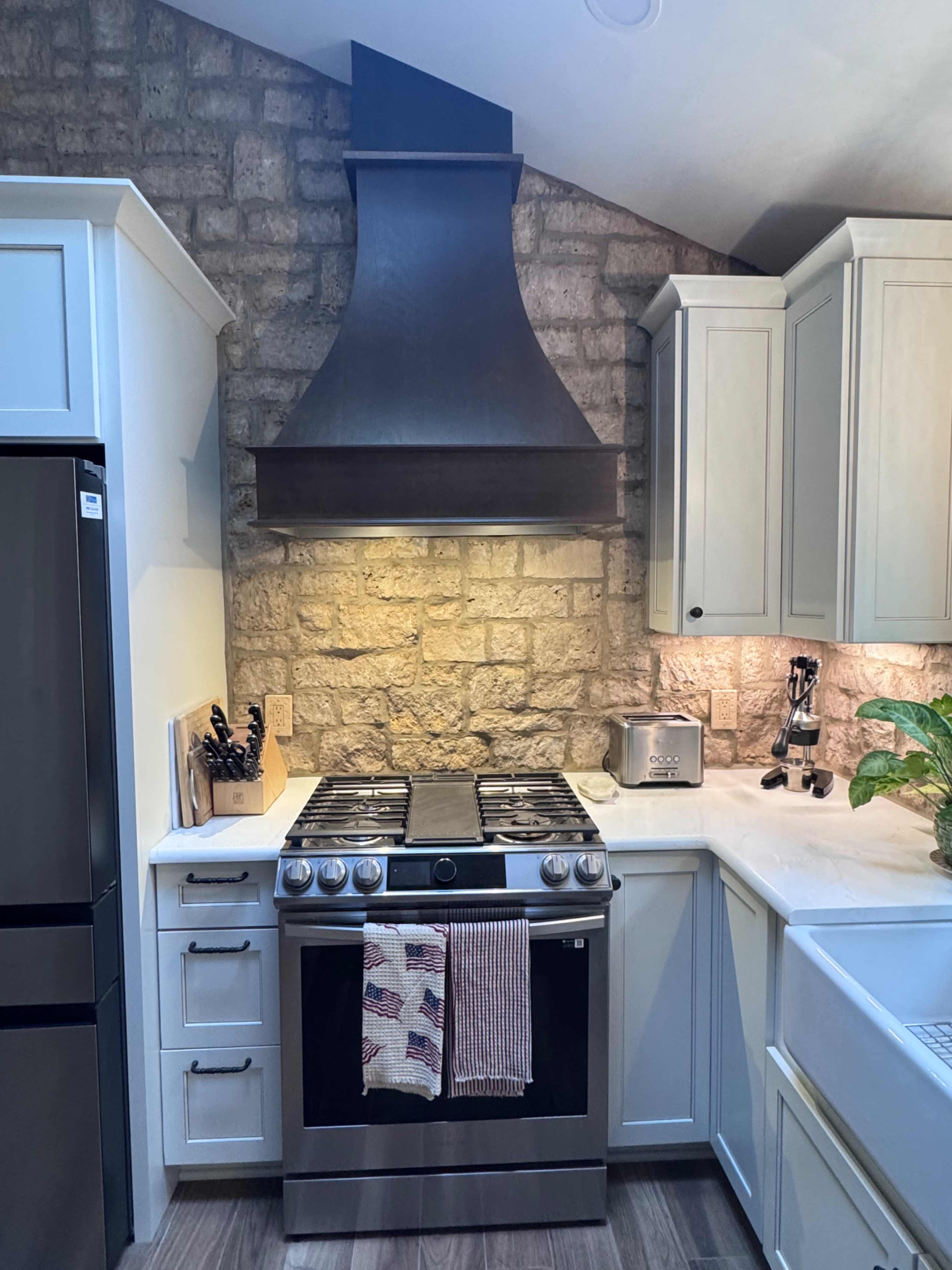 The image shows a modern kitchen featuring a dark metal range hood above a stainless steel gas stove, with stone accent wall and light-colored cabinetry.