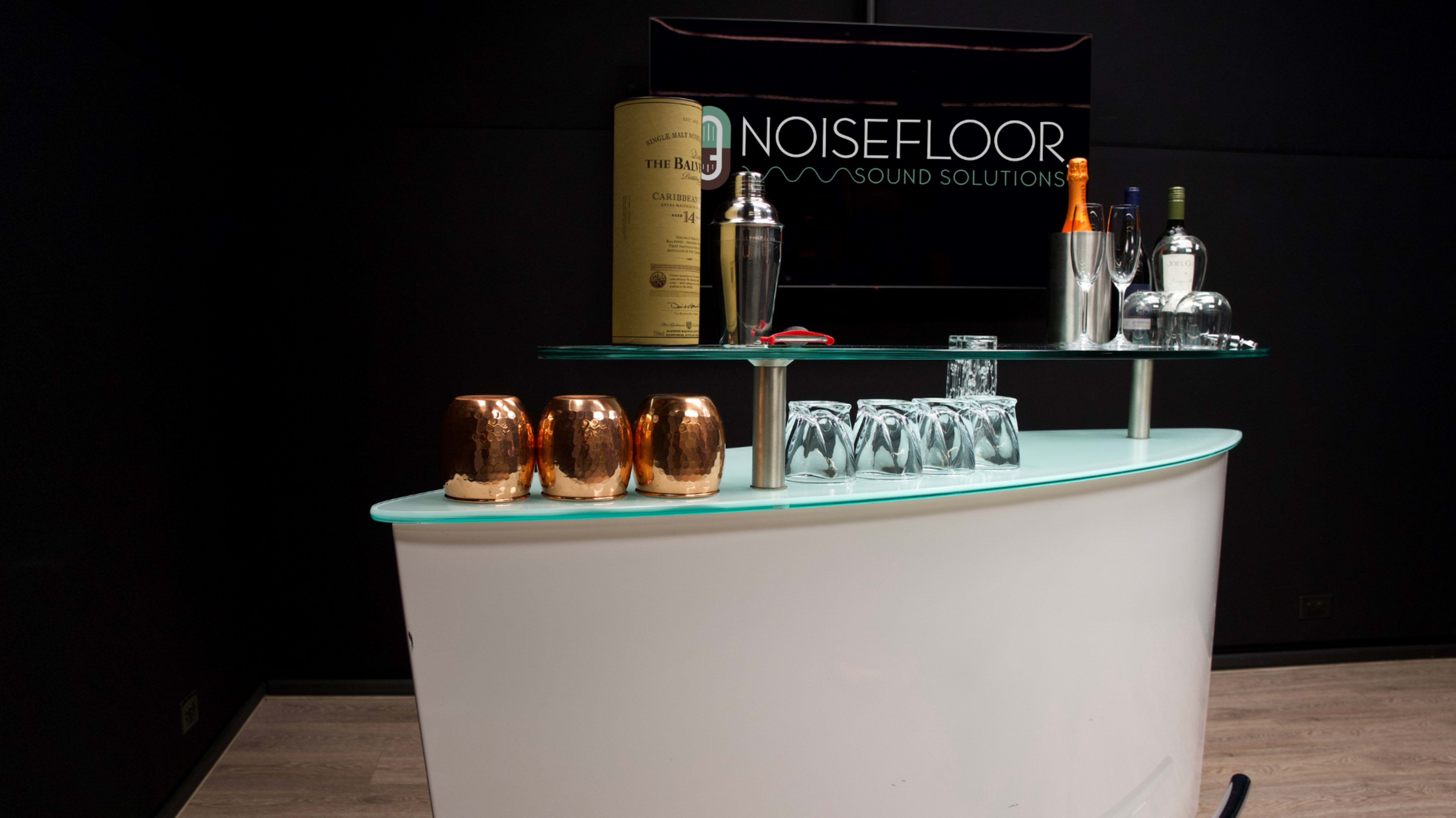 The image shows a sleek bar counter featuring a variety of bottles, a cocktail shaker, and copper mugs against a dark background with a company sign for "NoiseFloor Sound Solutions."
