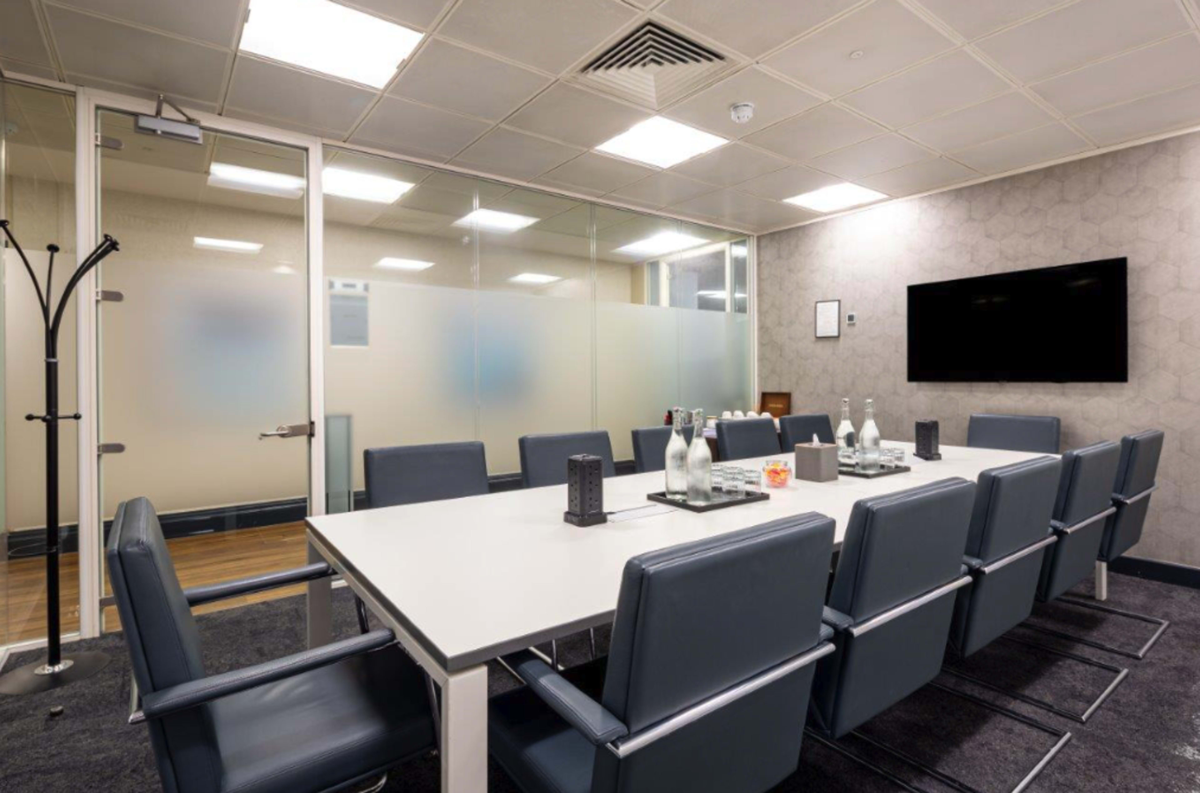A modern conference room with a long table surrounded by several leather chairs, glass walls, and a wall-mounted television.