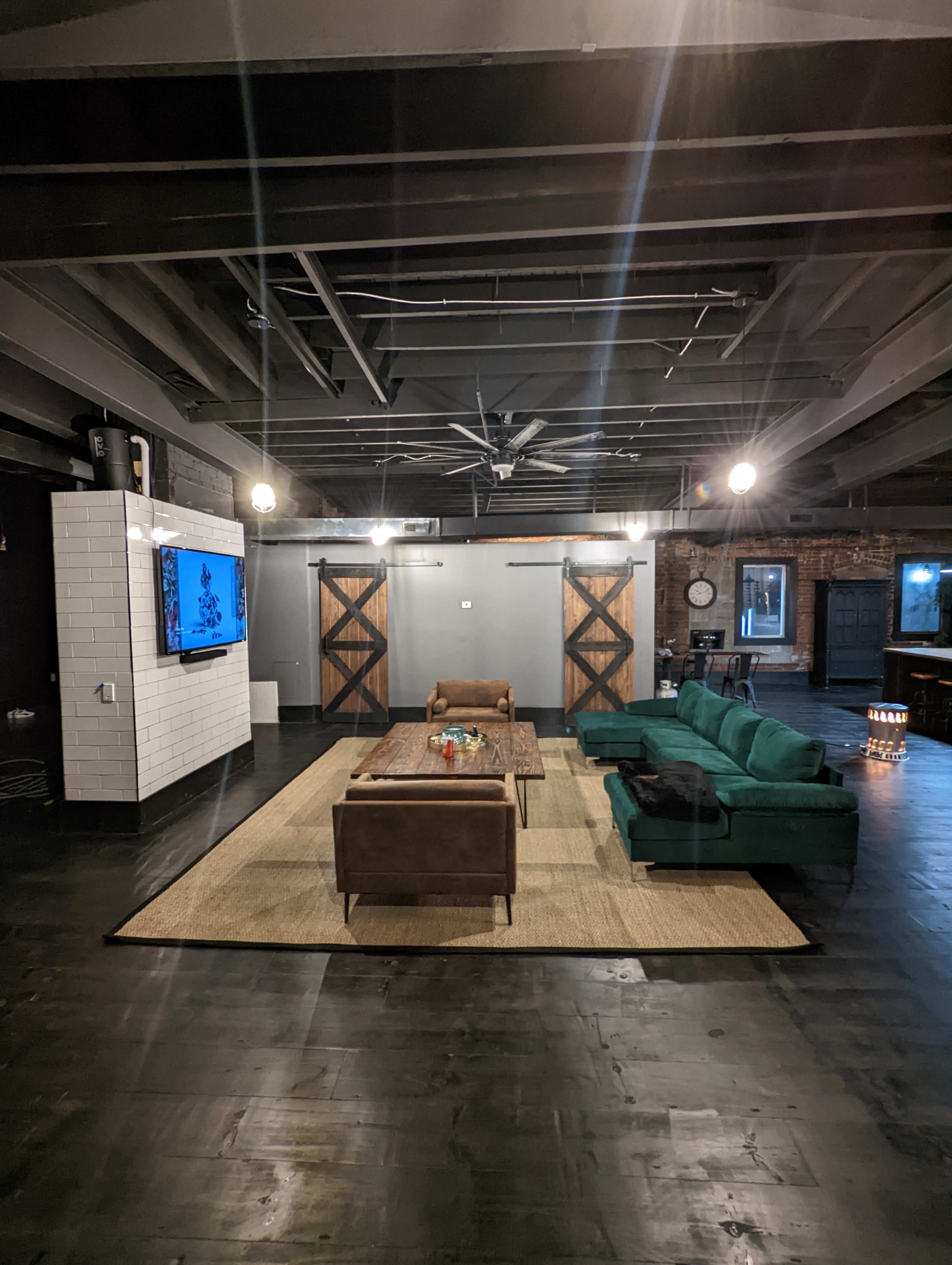 A spacious living area features a green sectional sofa and a wooden coffee table, with a large television mounted on a white wall and exposed brick accents.