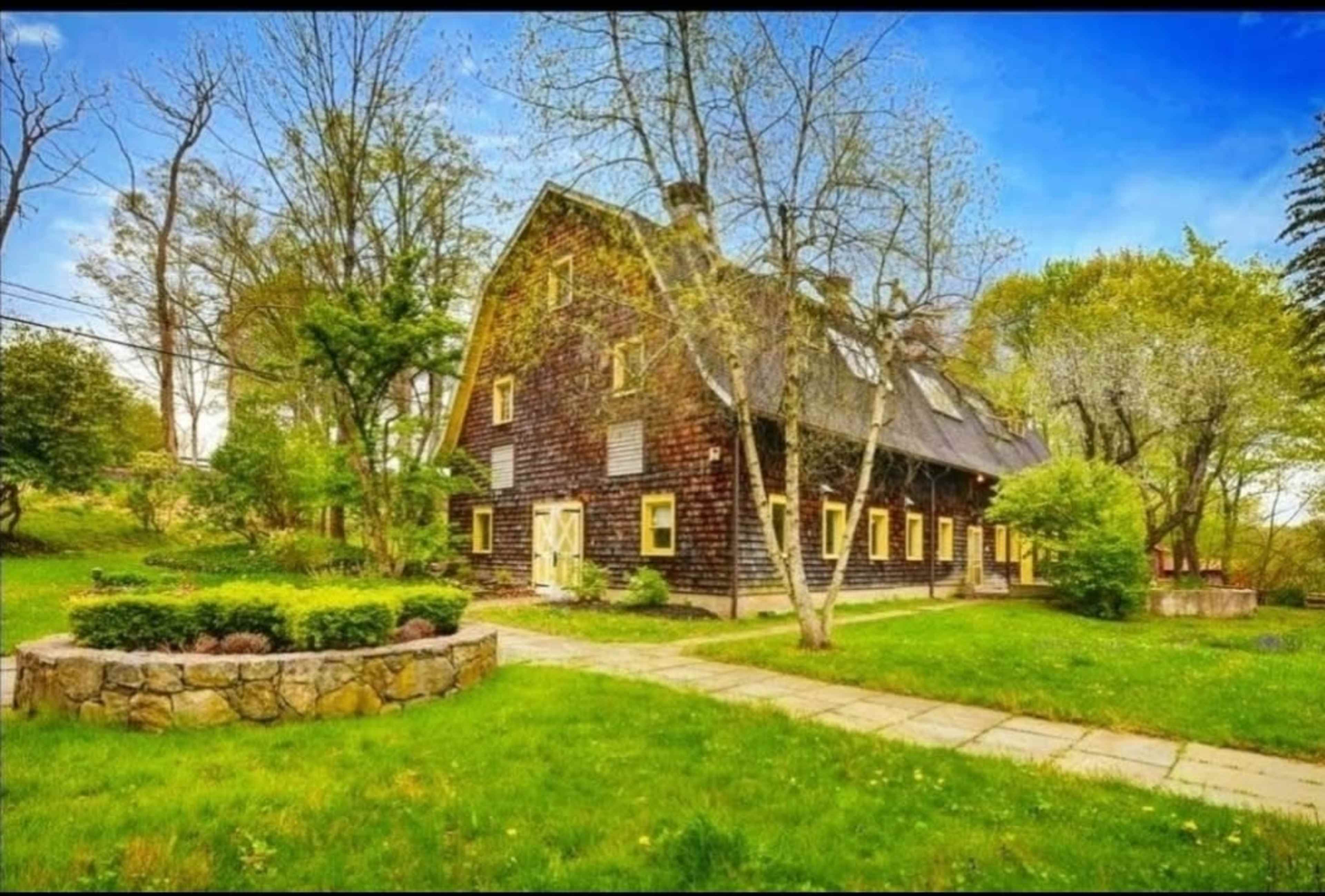 A large wooden house with a steep roof, surrounded by green grass and trees, with a stone pathway leading to it.