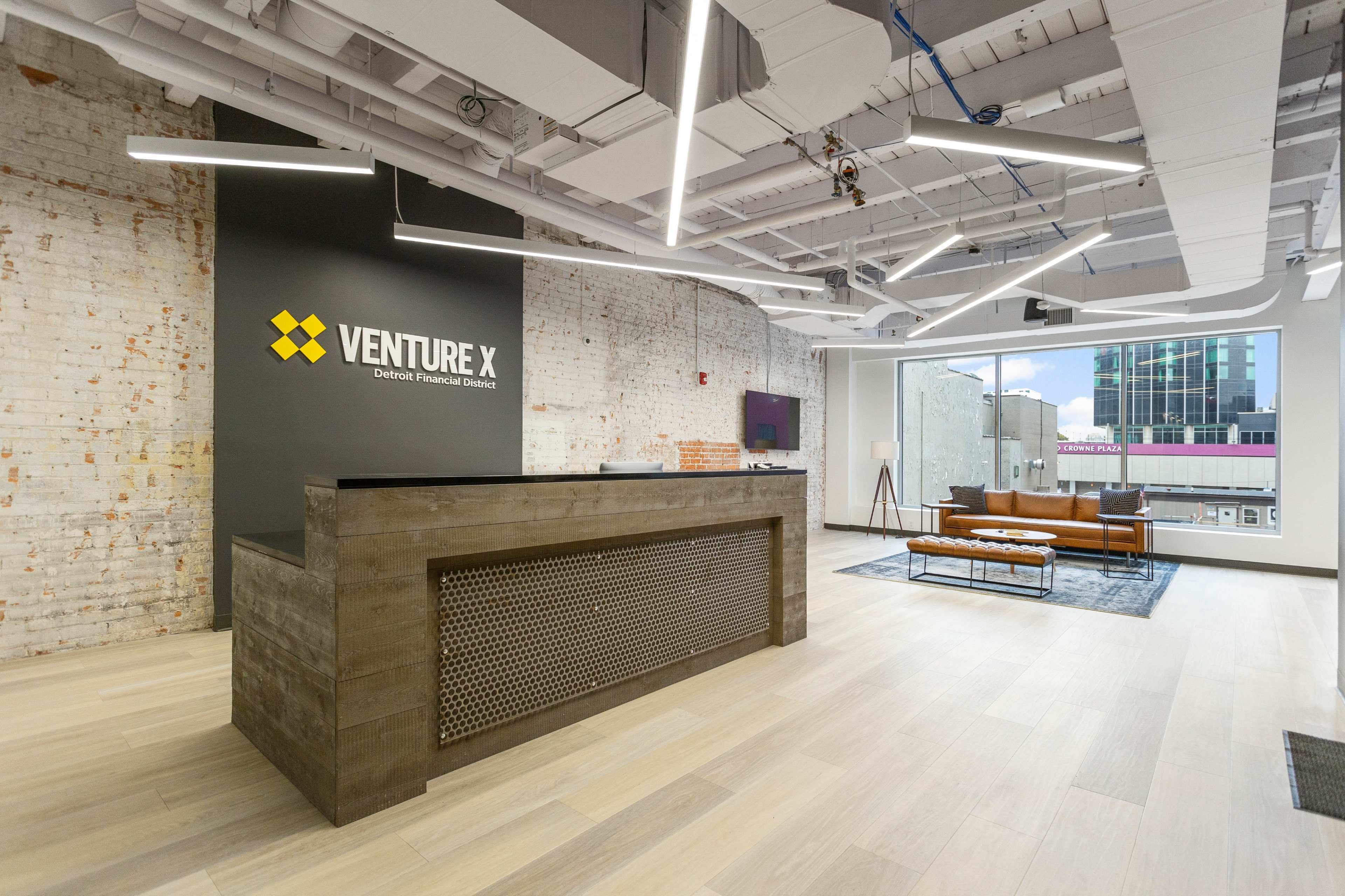 A modern office reception area with a wooden front desk featuring the logo "VENTURE X" on a gray wall, surrounded by large windows and a seating area with a brown sofa.