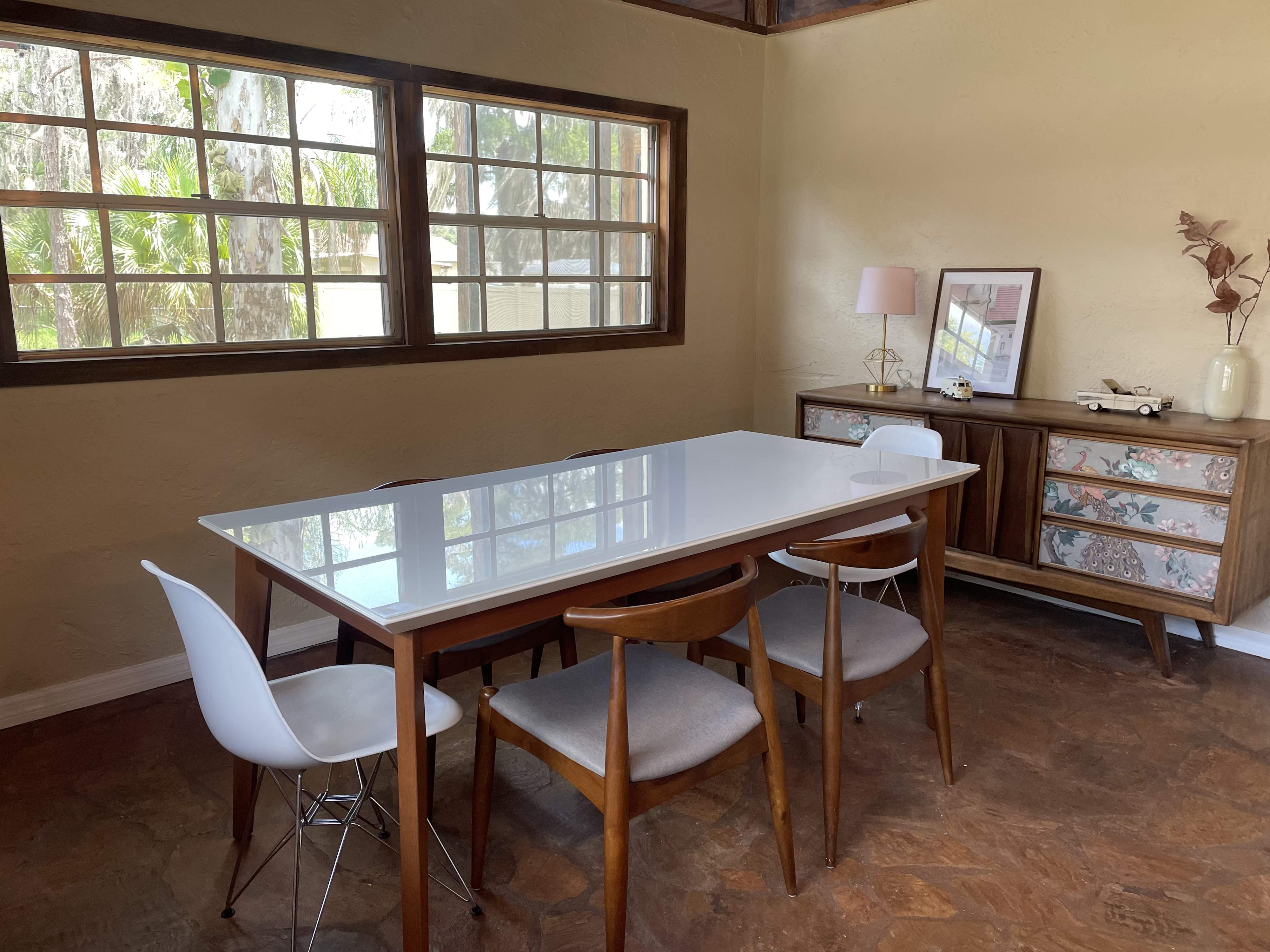 A dining area features a white table surrounded by four wooden chairs, with a cabinet and a window displaying greenery outside.
