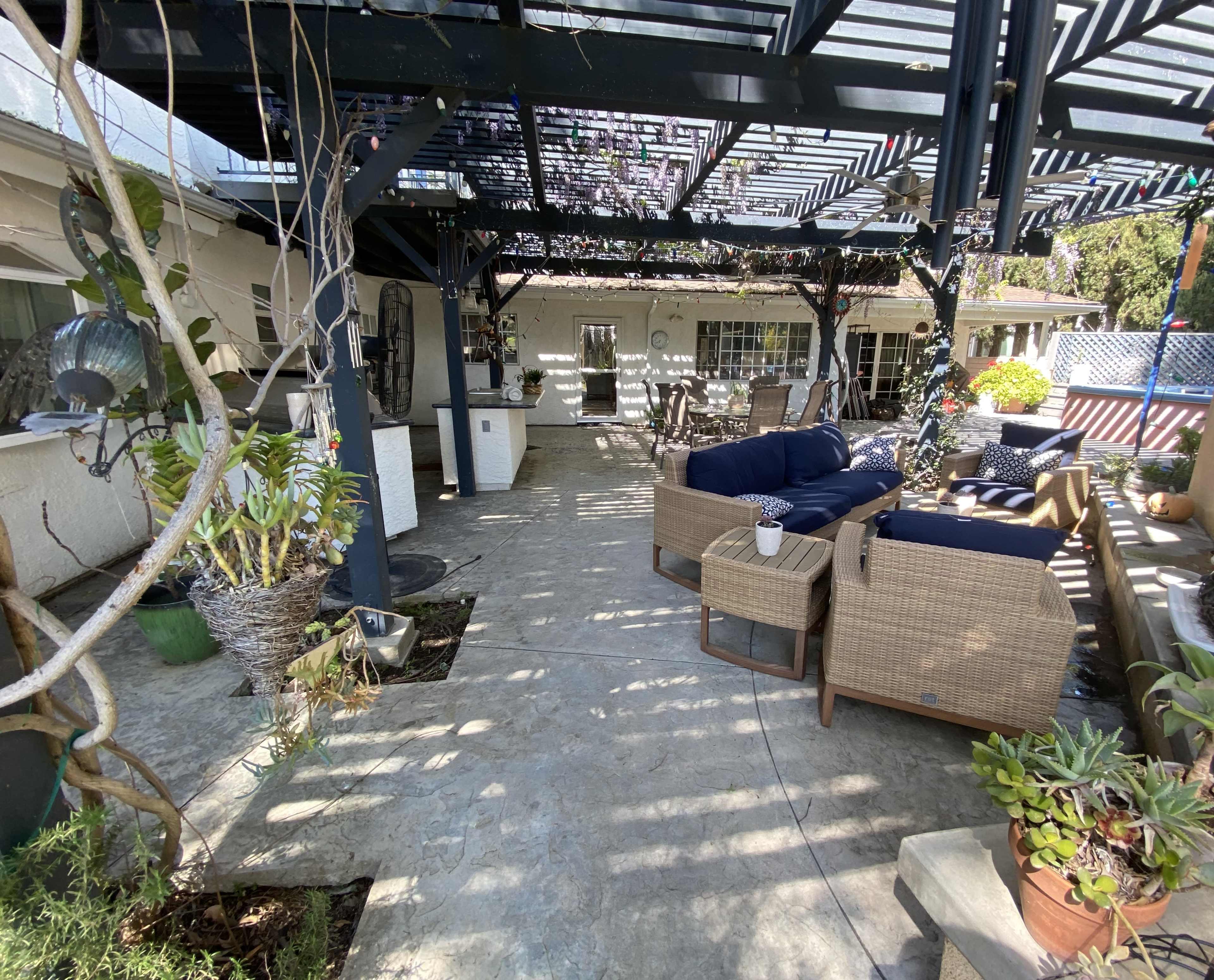 The image shows a covered outdoor patio area with seating and potted plants, featuring a concrete floor and a lattice roof.