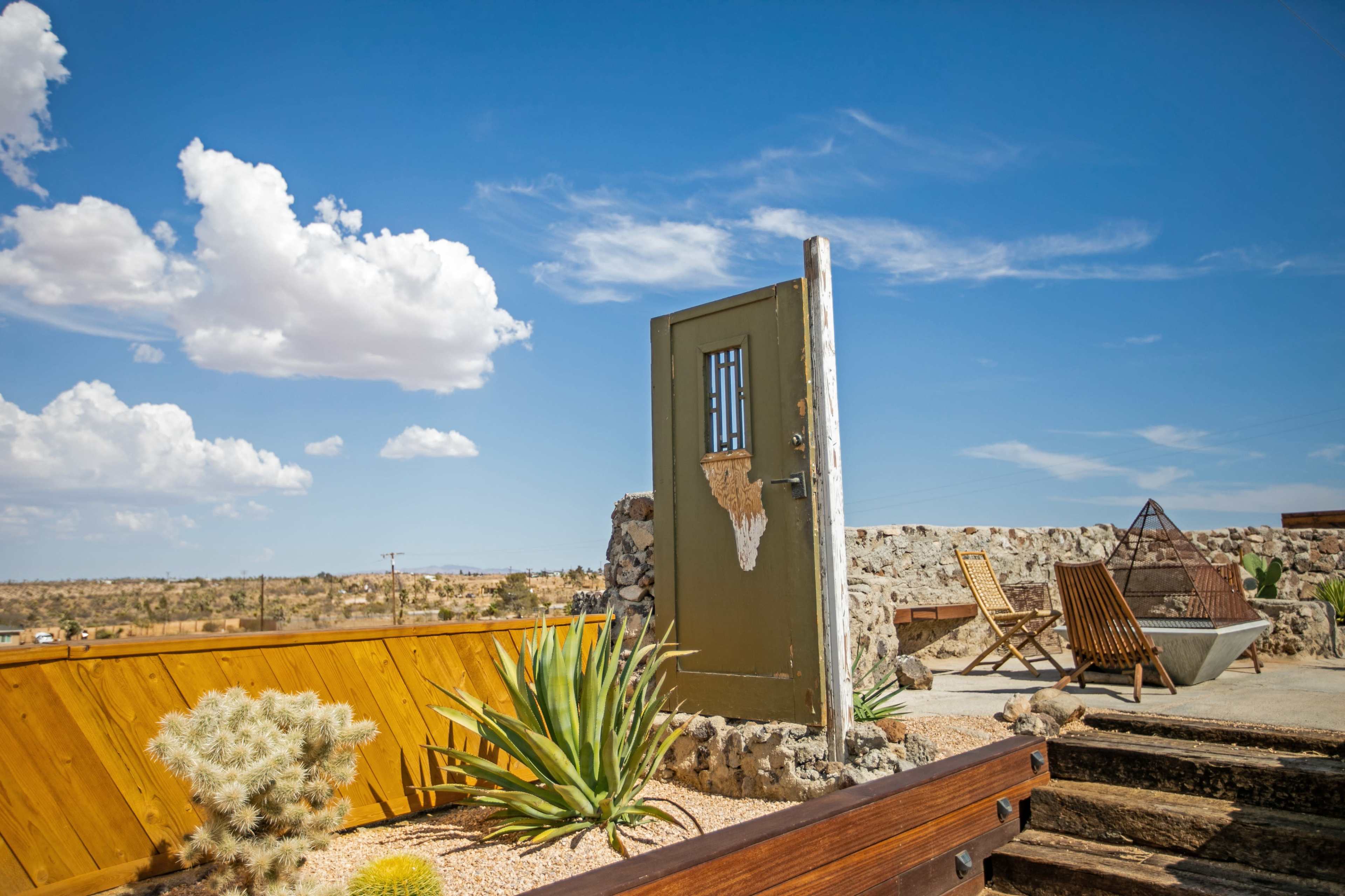 A weathered green door stands alone on a stone structure with desert plants and wooden furniture under a blue sky with scattered clouds.