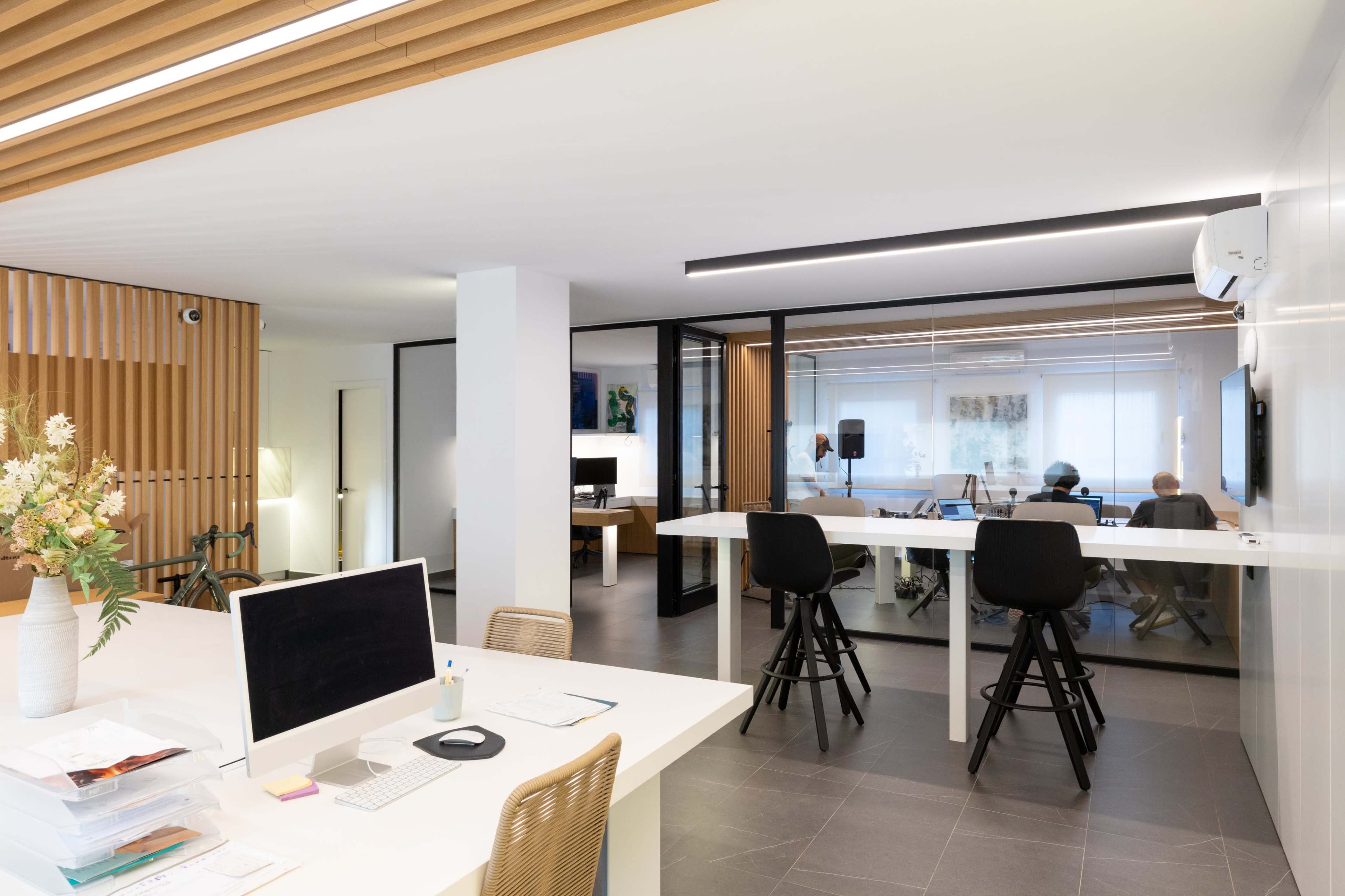 An open office space features a reception area with a computer and paperwork, and a glass-walled meeting room where two people are seated.