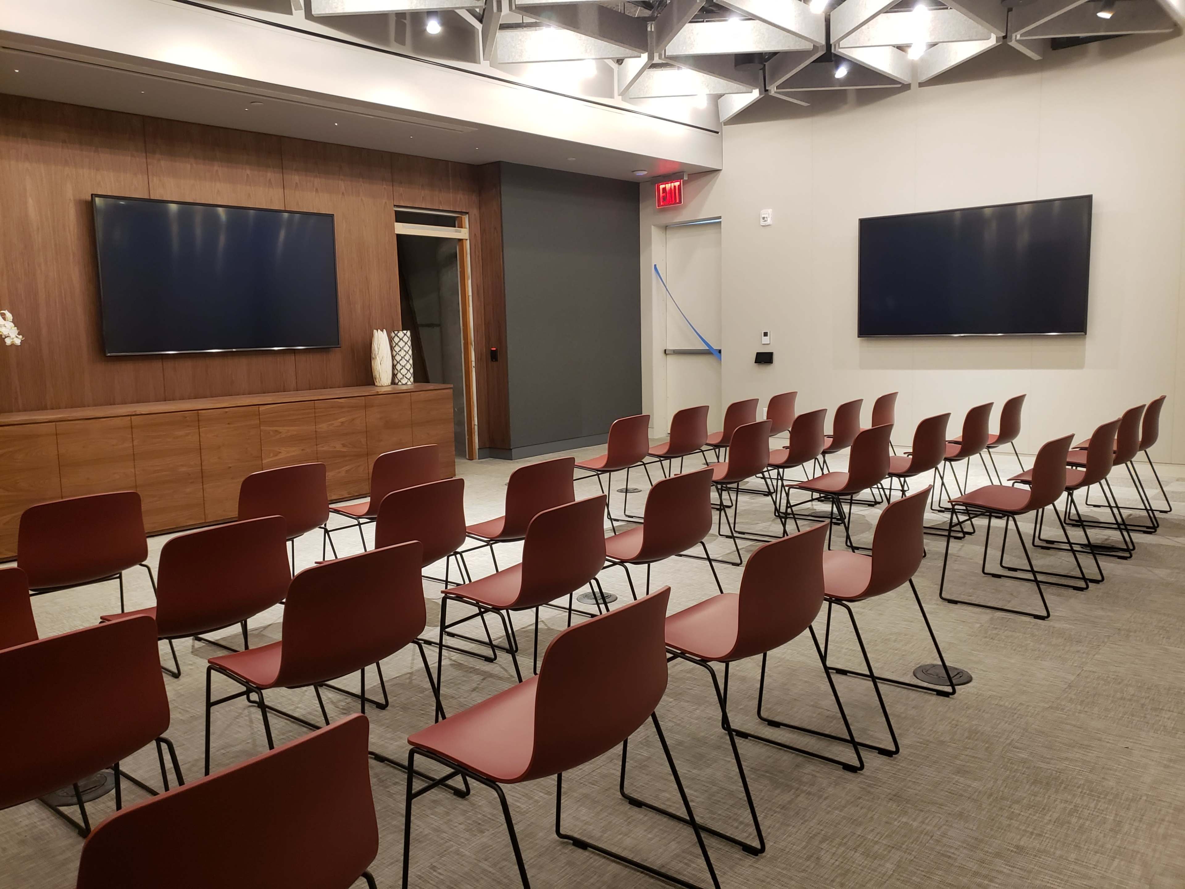 A meeting room with rows of red chairs facing two wall-mounted screens.