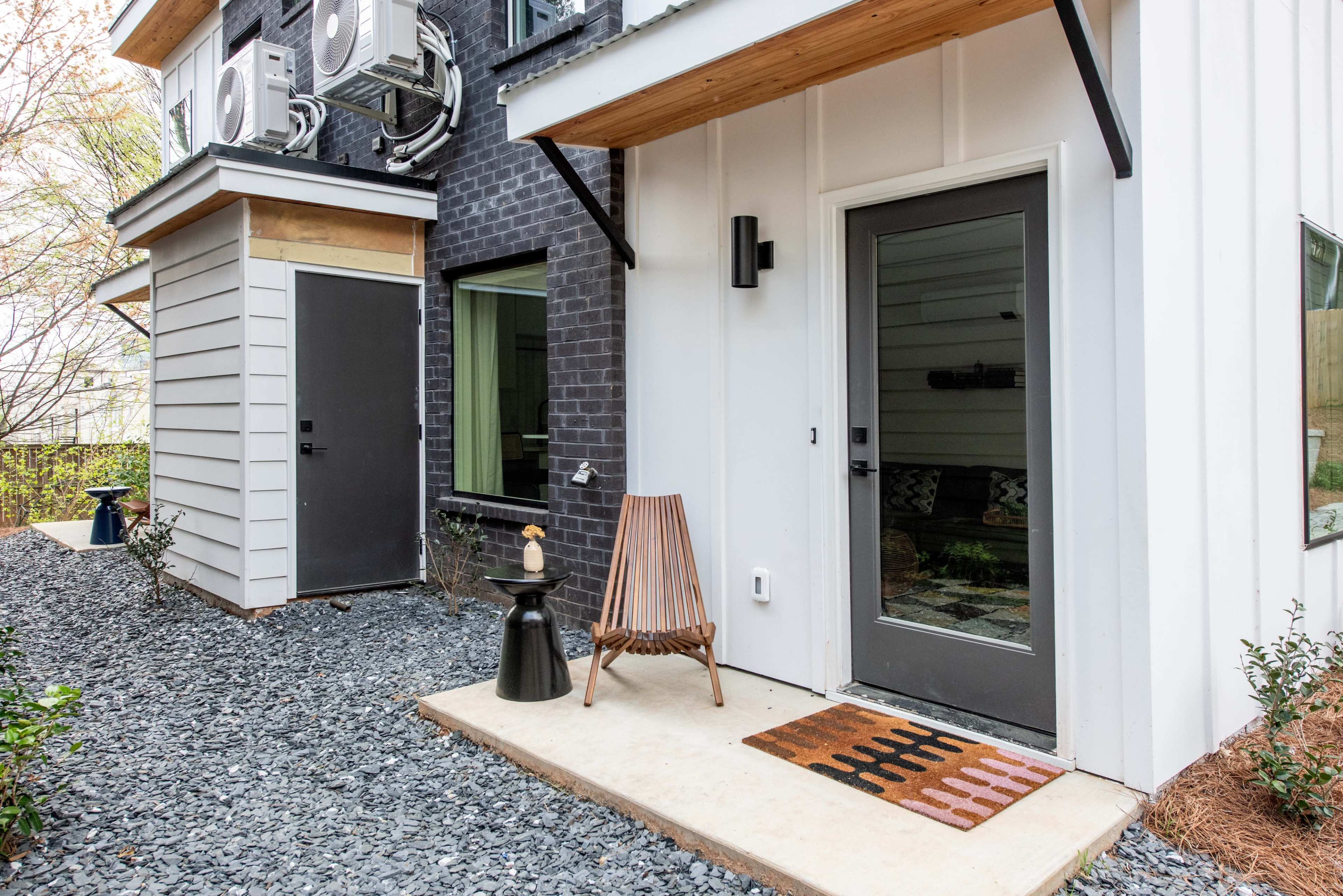 The image shows a modern home entrance featuring a wooden chair, a patterned doormat, and air conditioning units mounted on the wall.