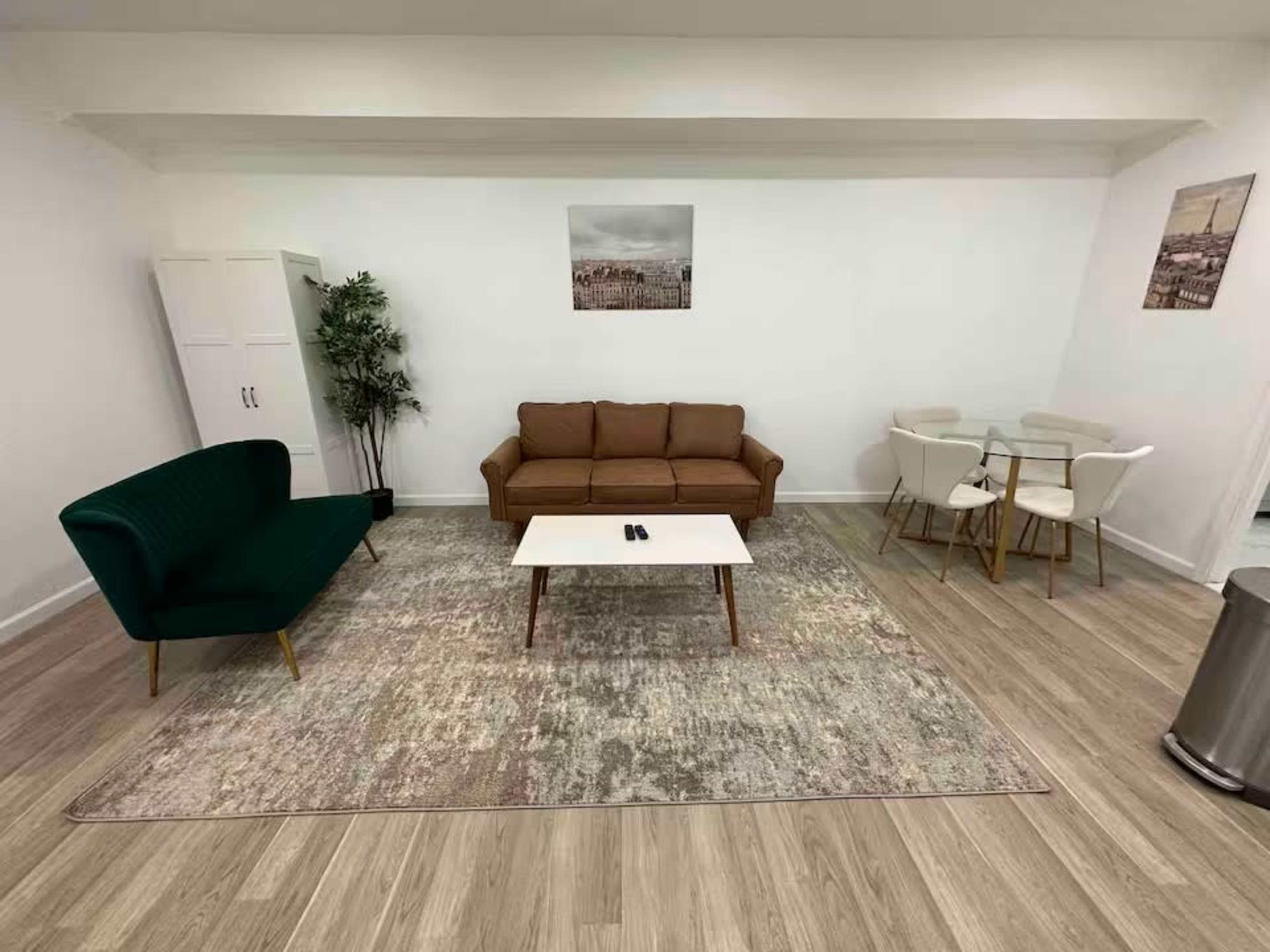 The image shows a minimalist living space featuring a green upholstered sofa, a brown couch, a glass dining table with white chairs, and a patterned area rug.