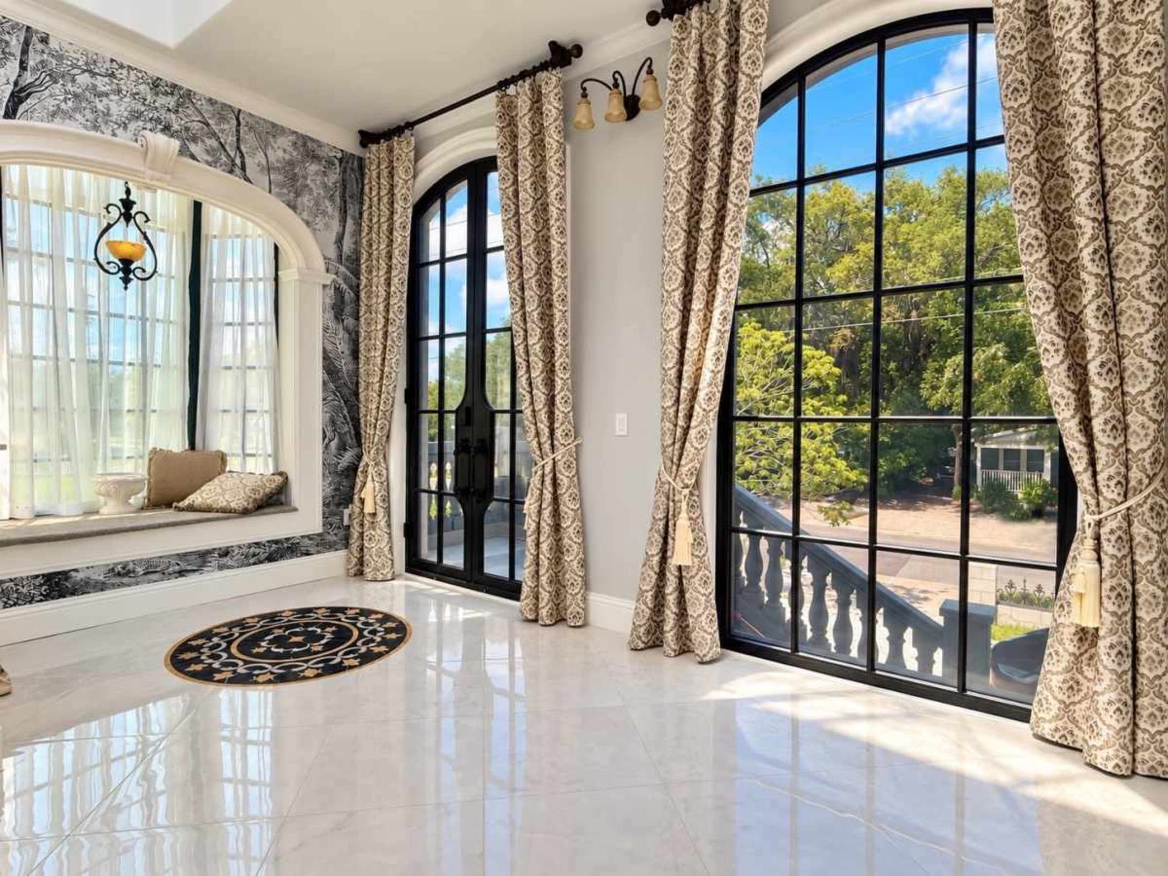 The room features large arched windows with decorative curtains, a cozy window seat, and a round rug against a backdrop of intricate wallpaper.