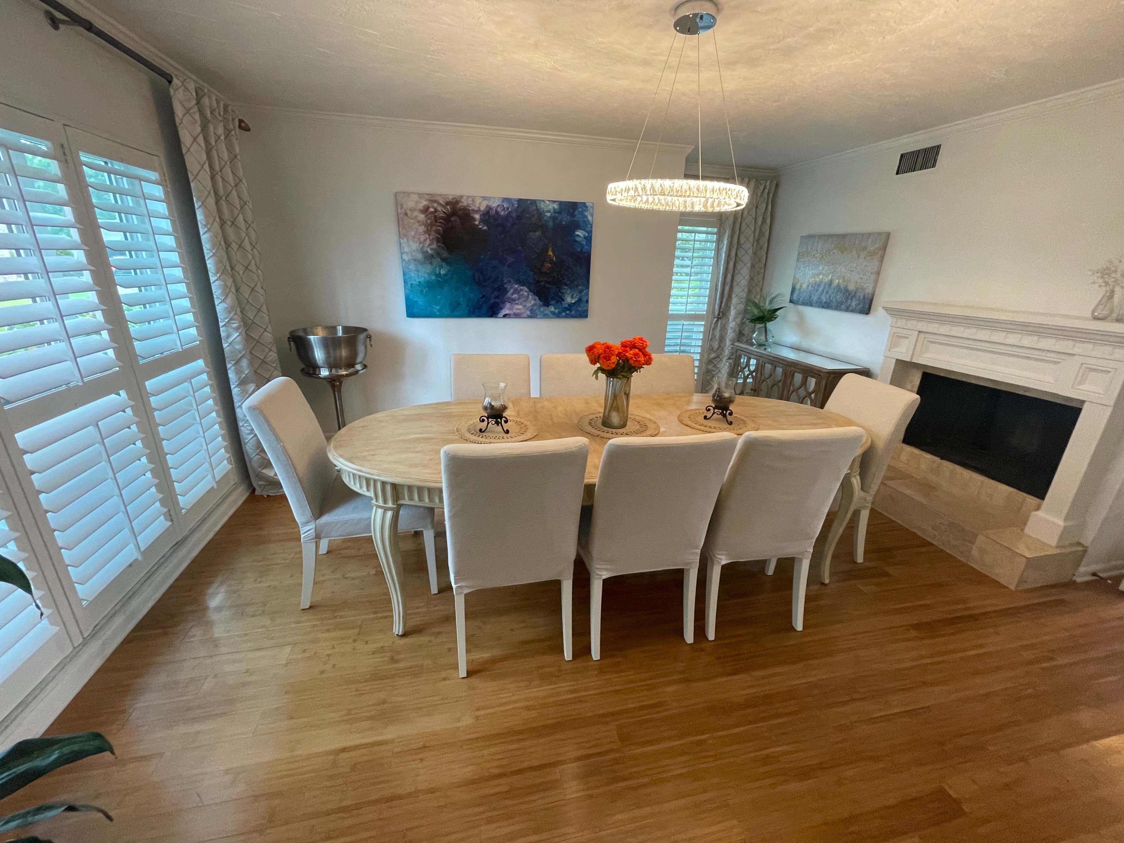A round wooden dining table surrounded by eight white chairs is set in a well-lit room featuring a fireplace and artwork on the walls.