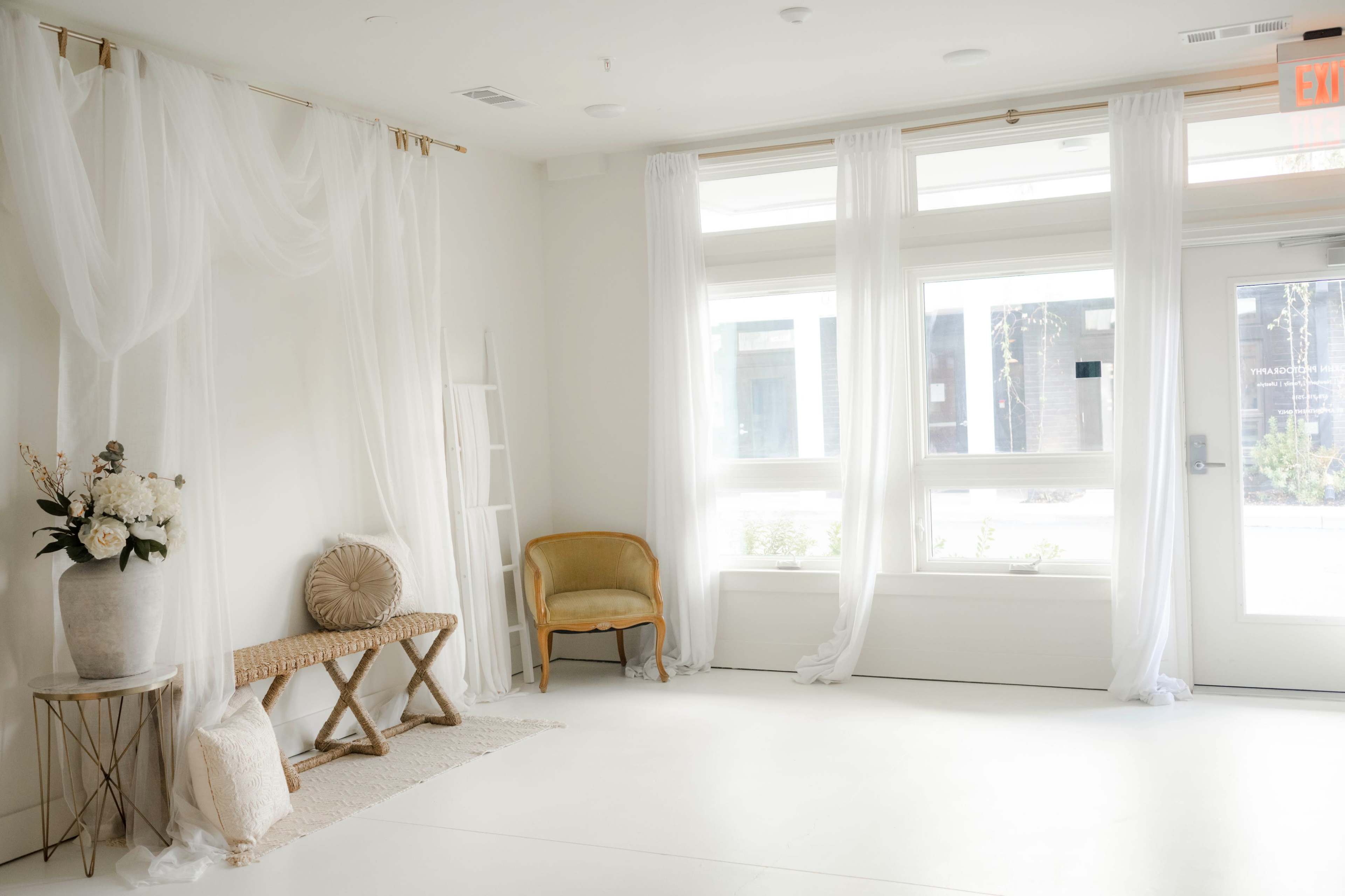 The scene features a bright and airy indoor space with sheer white drapes, a wicker chair, a large vase of flowers, and a simple woven rug on a light-colored floor.