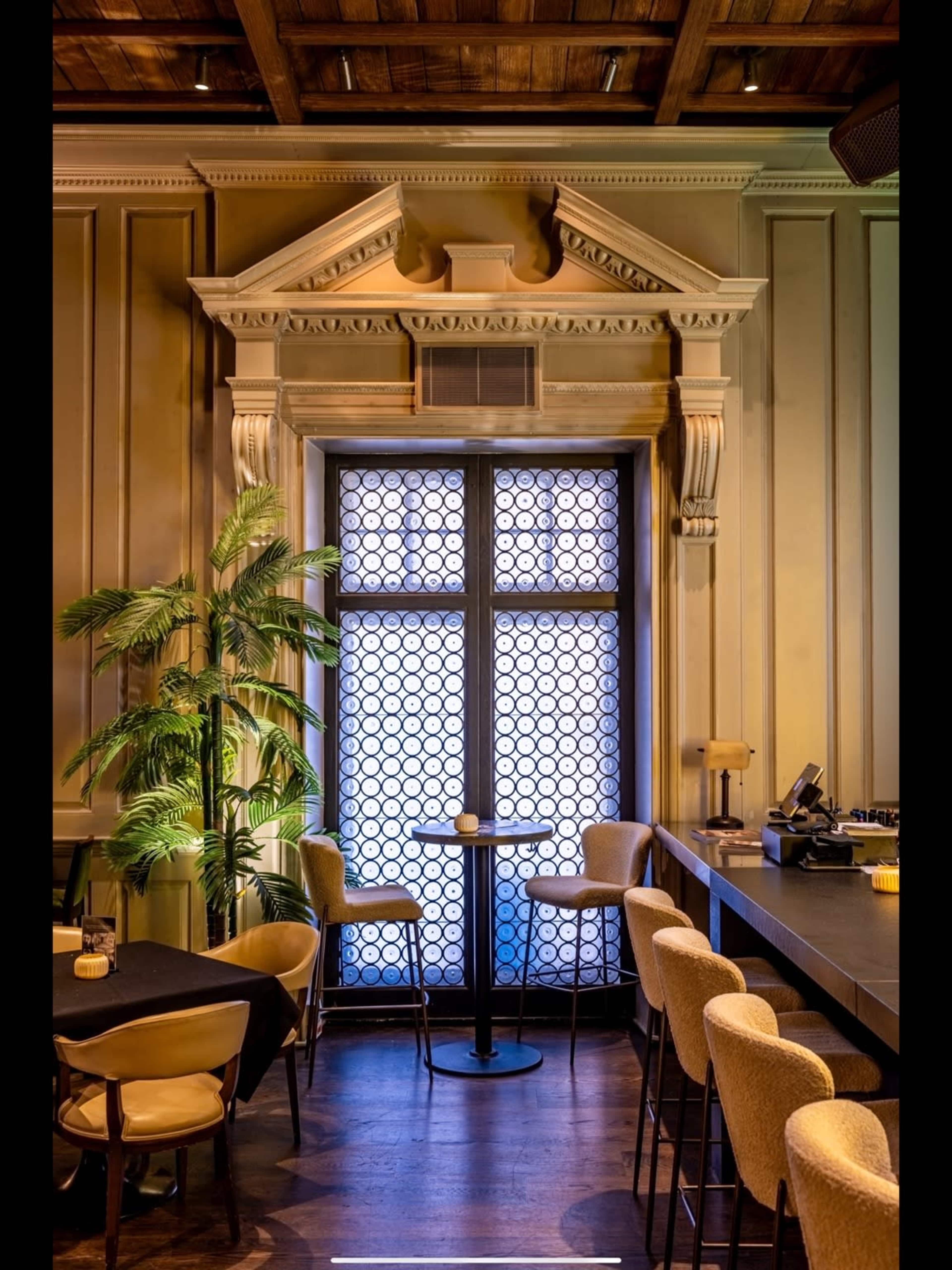 The scene features an elegant interior of a restaurant with a window adorned by a decorative architectural feature, a few bar stools, and a tall plant beside the table.