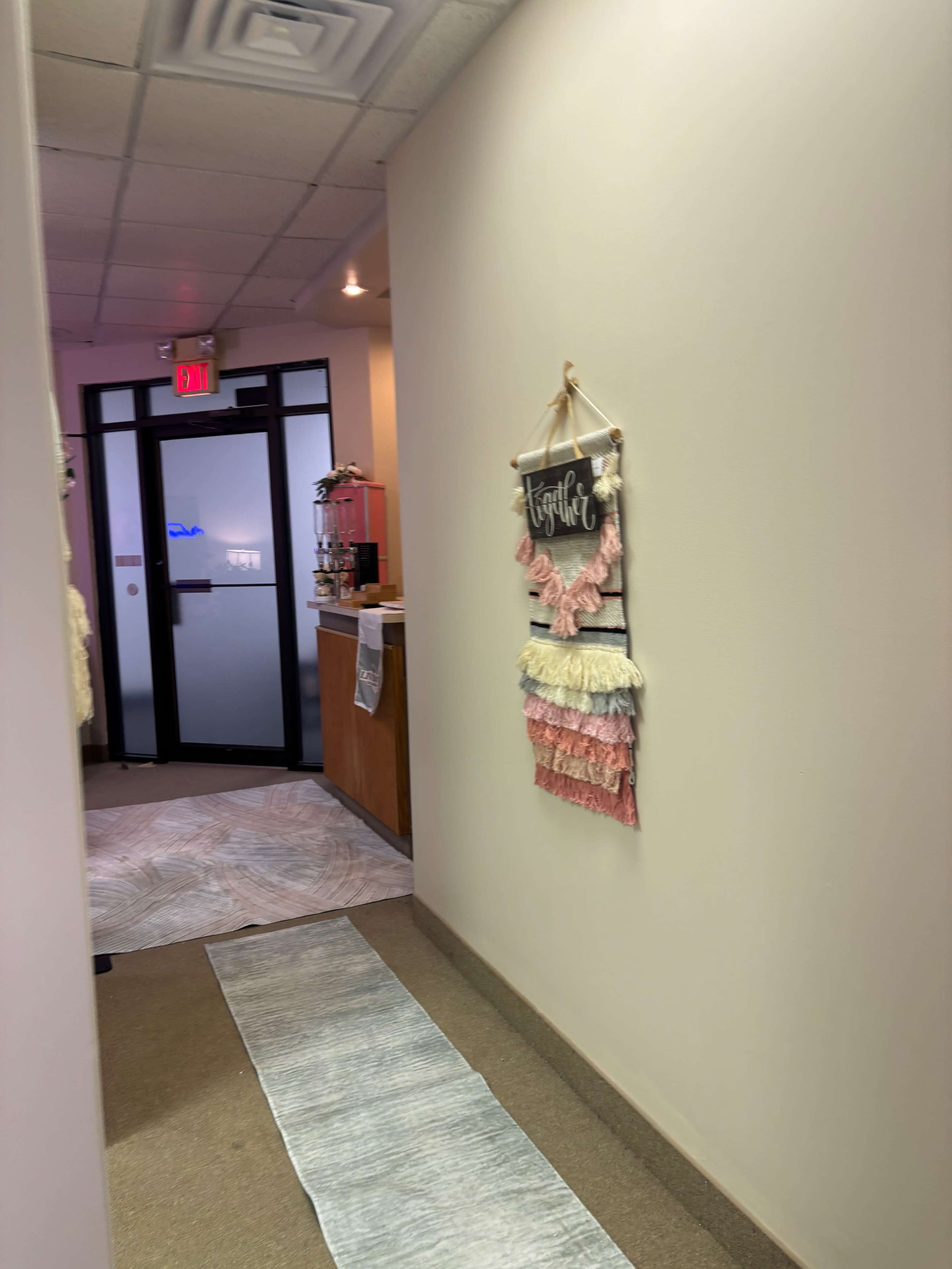 The image shows a narrow hallway leading to an office with a decorative wall hanging on the left and a reception area visible at the end.
