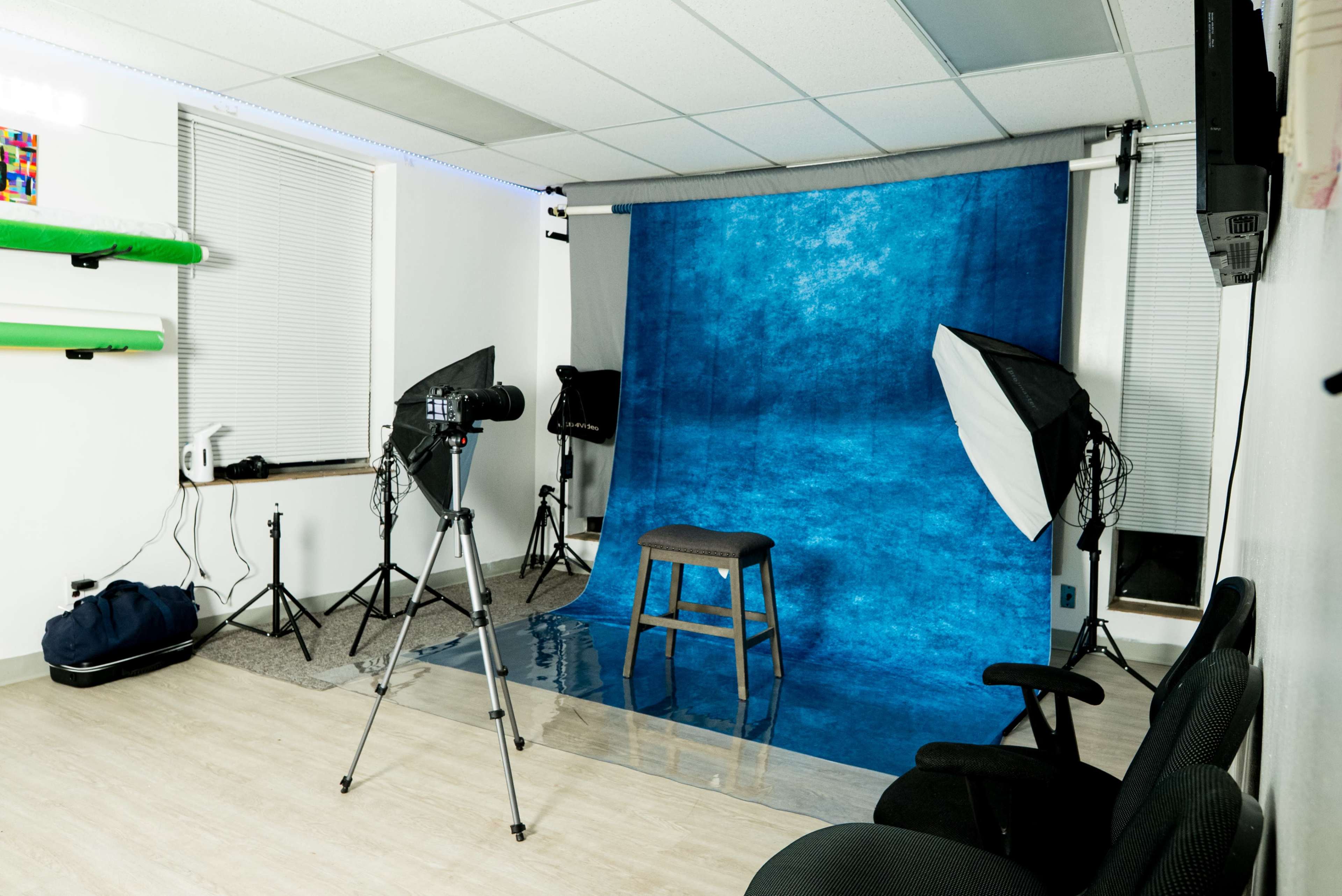 A photography studio features a blue backdrop, a stool, lighting equipment, and a camera on a tripod.