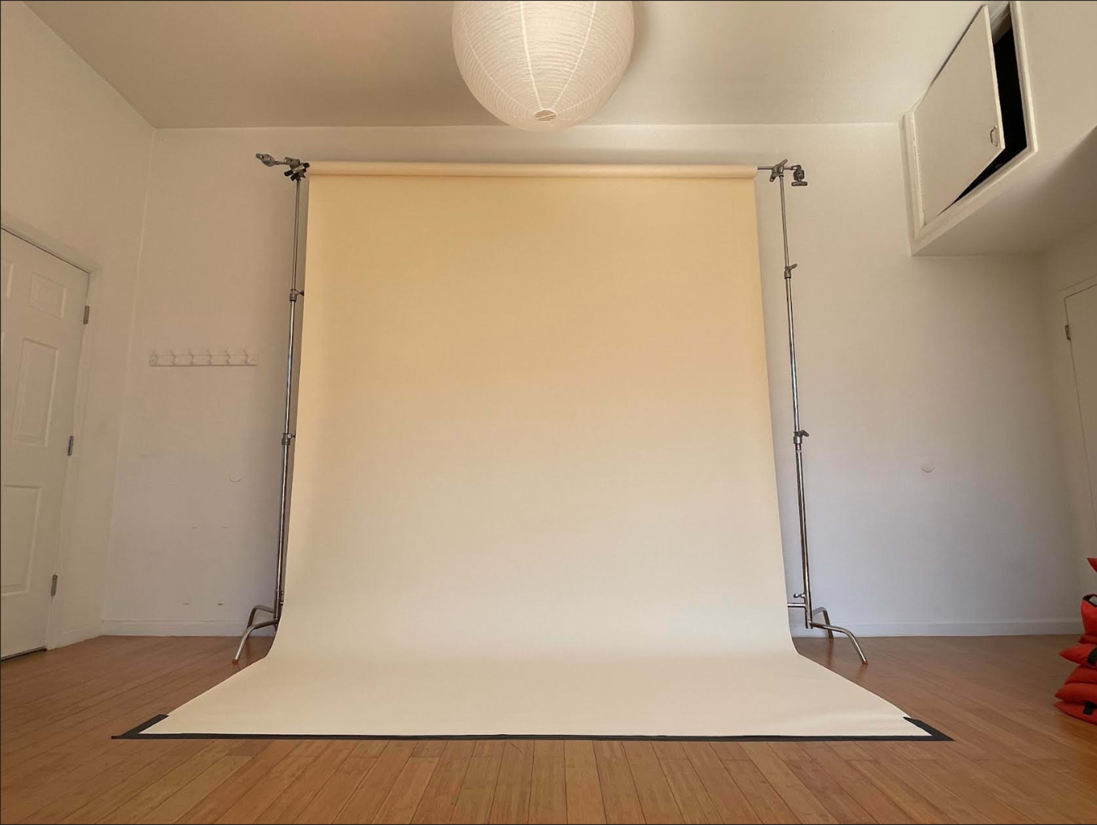 A minimalist photography studio with a beige backdrop set on a wooden floor, illuminated by a pendant light.