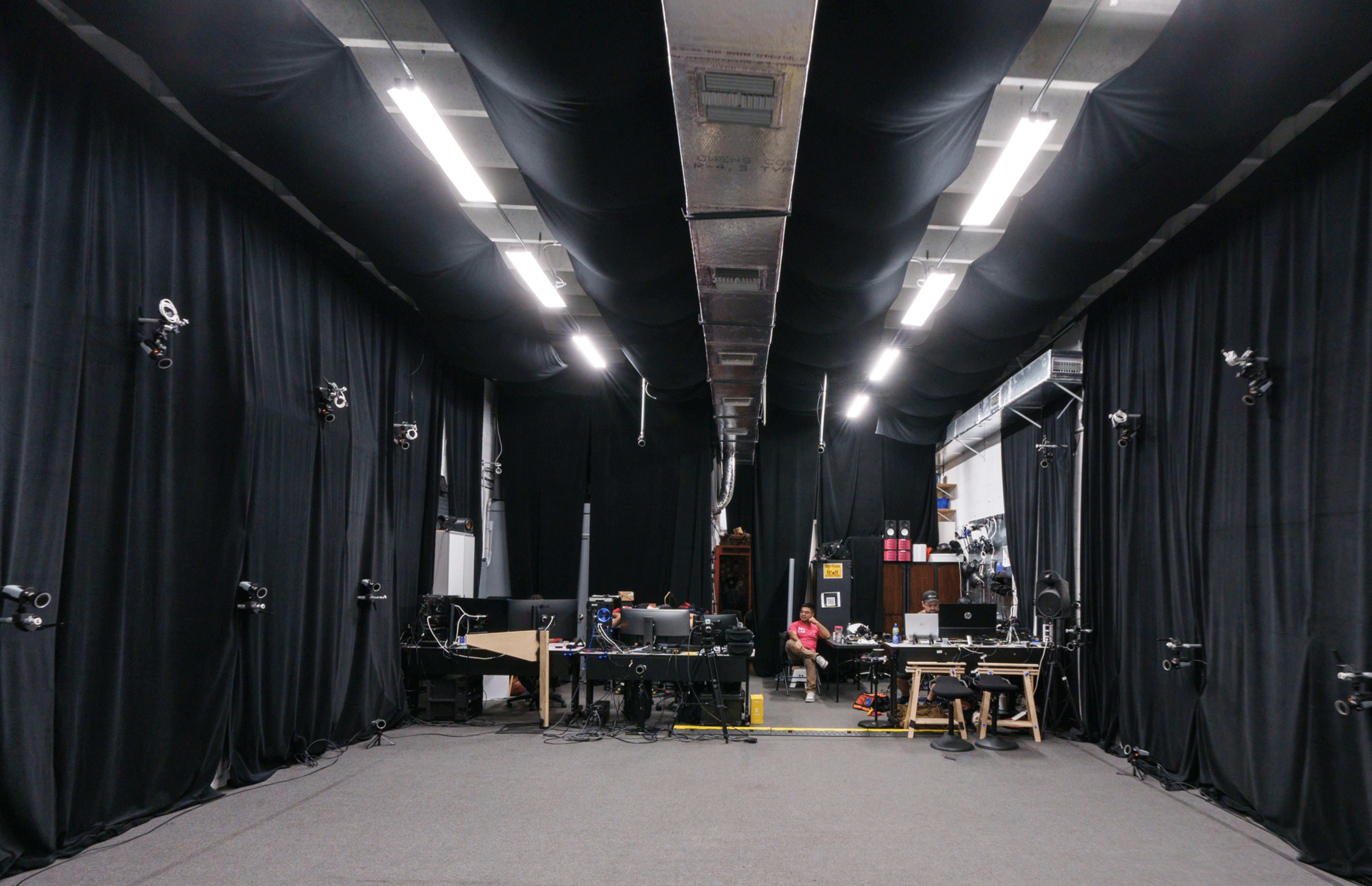 The image shows a large indoor space with black curtains lining the walls and various equipment arranged in the center, including tables and cameras.