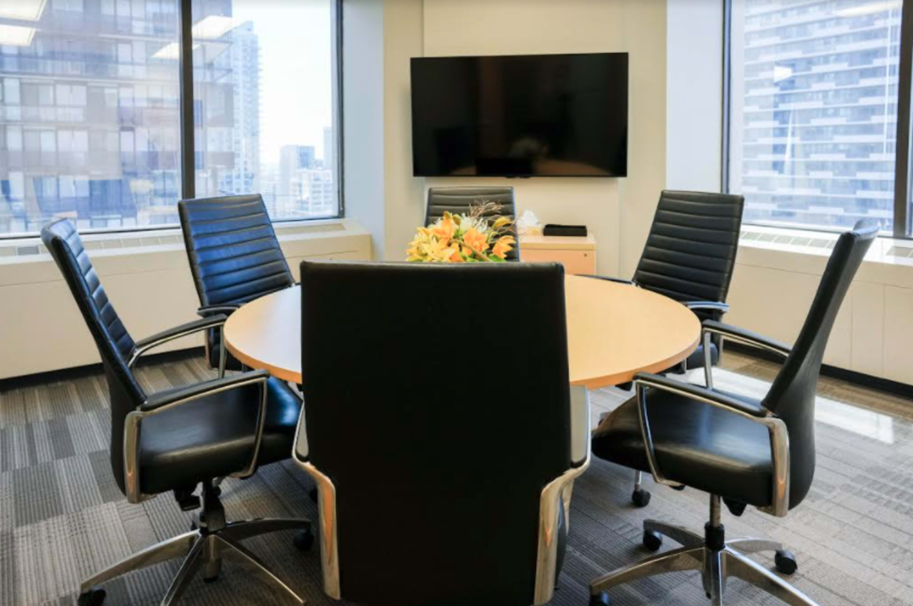 A conference room features a round table surrounded by five black chairs and a television mounted on the wall.