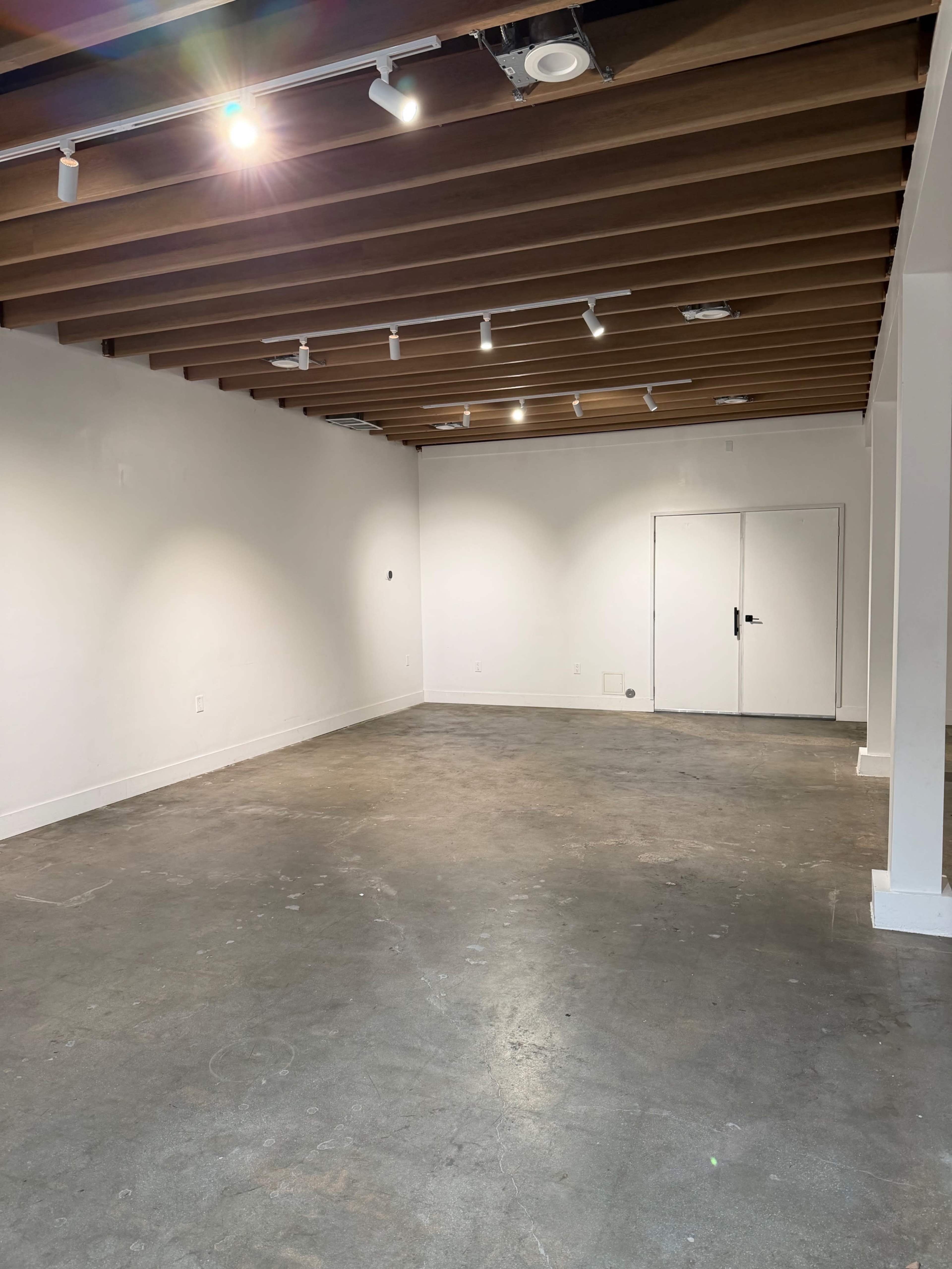 The image shows a spacious, empty room with concrete flooring and wooden ceiling beams, featuring overhead lighting and a closed double door at one end.