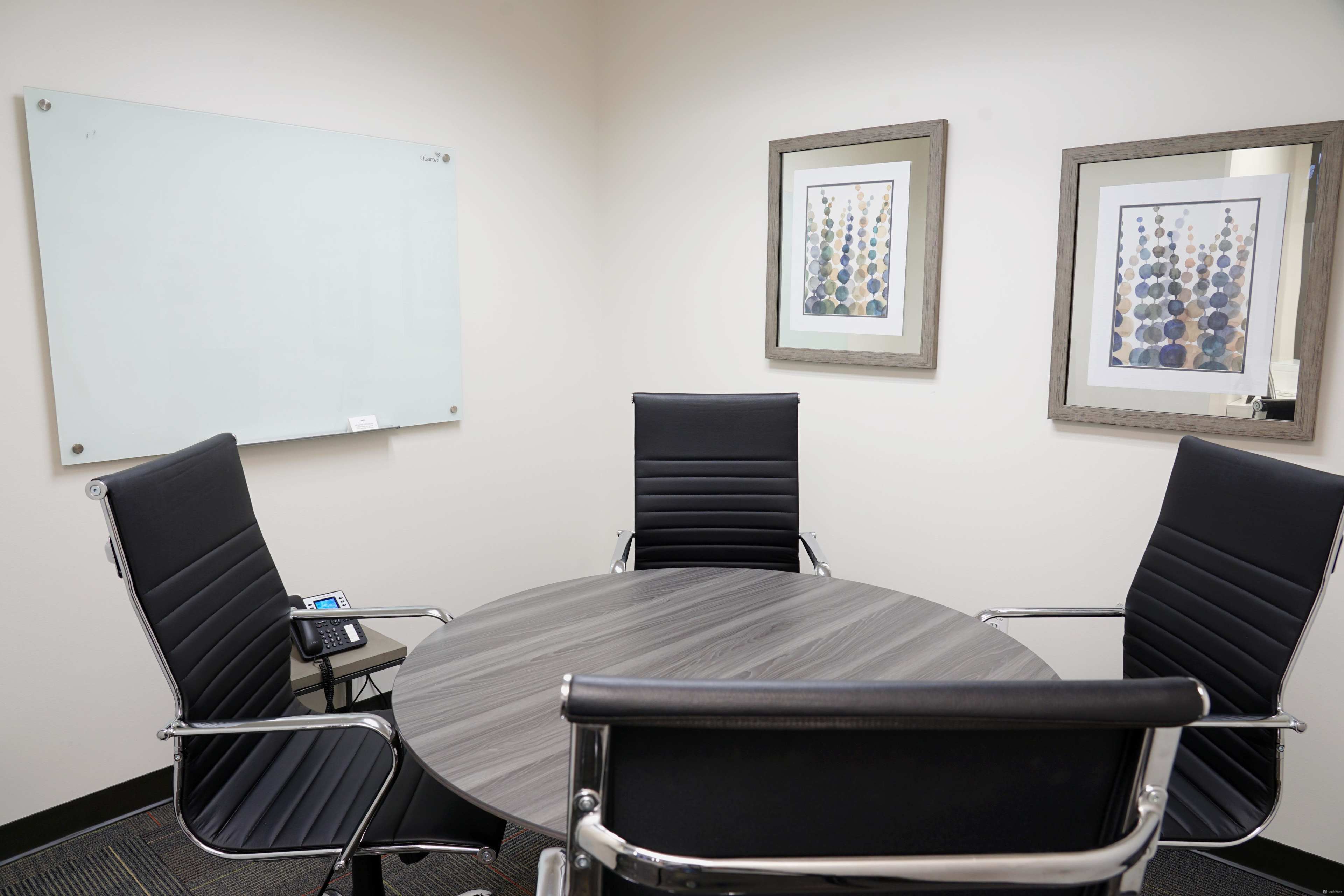 A small conference room features a circular table surrounded by four black chairs, with a whiteboard on one wall and two framed artworks nearby.