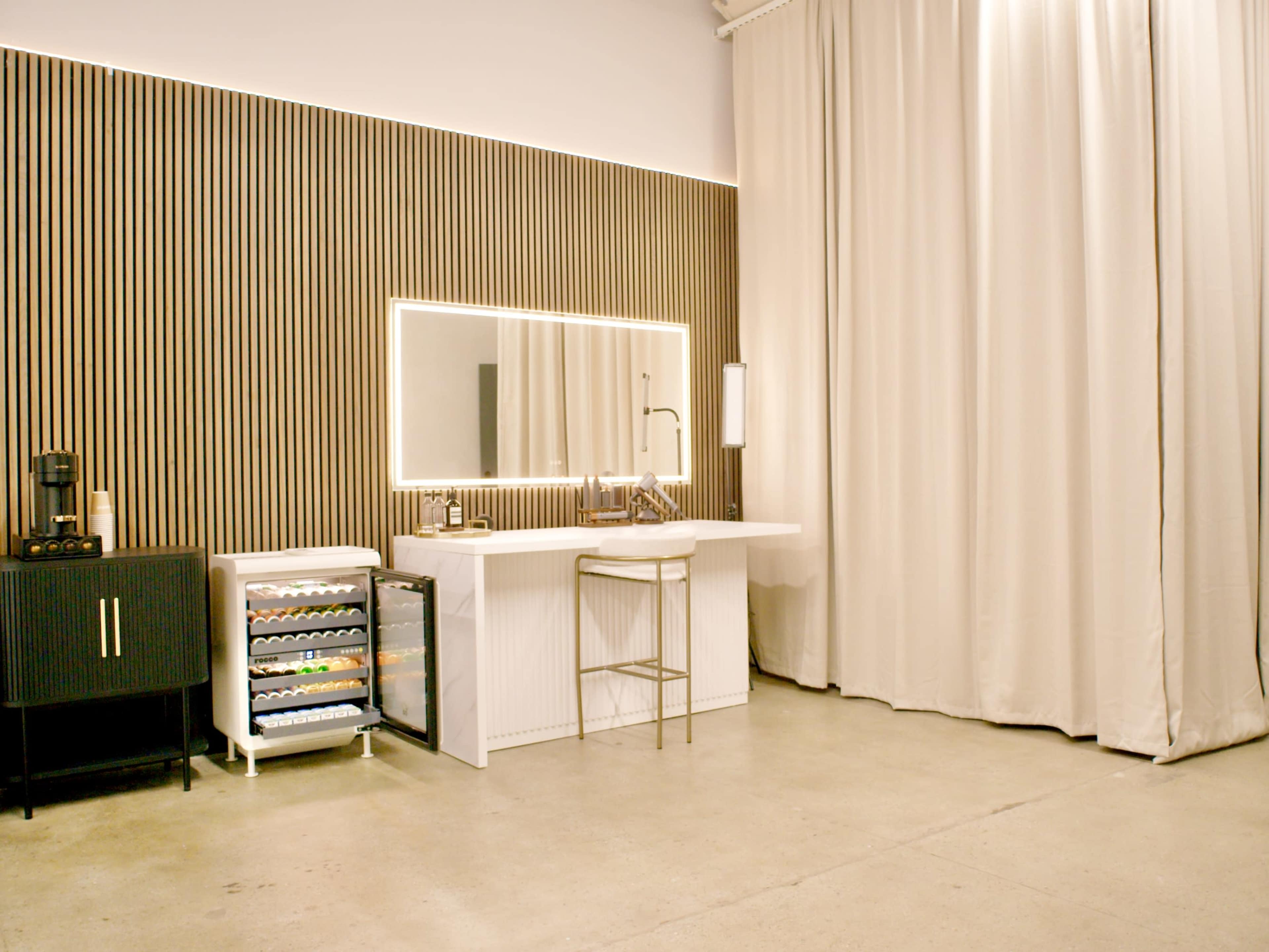 The image shows a modern vanity area with a large mirror, a white countertop, a bar stool, and a small refrigerator, all set against a backdrop of wooden paneling and a curtain.