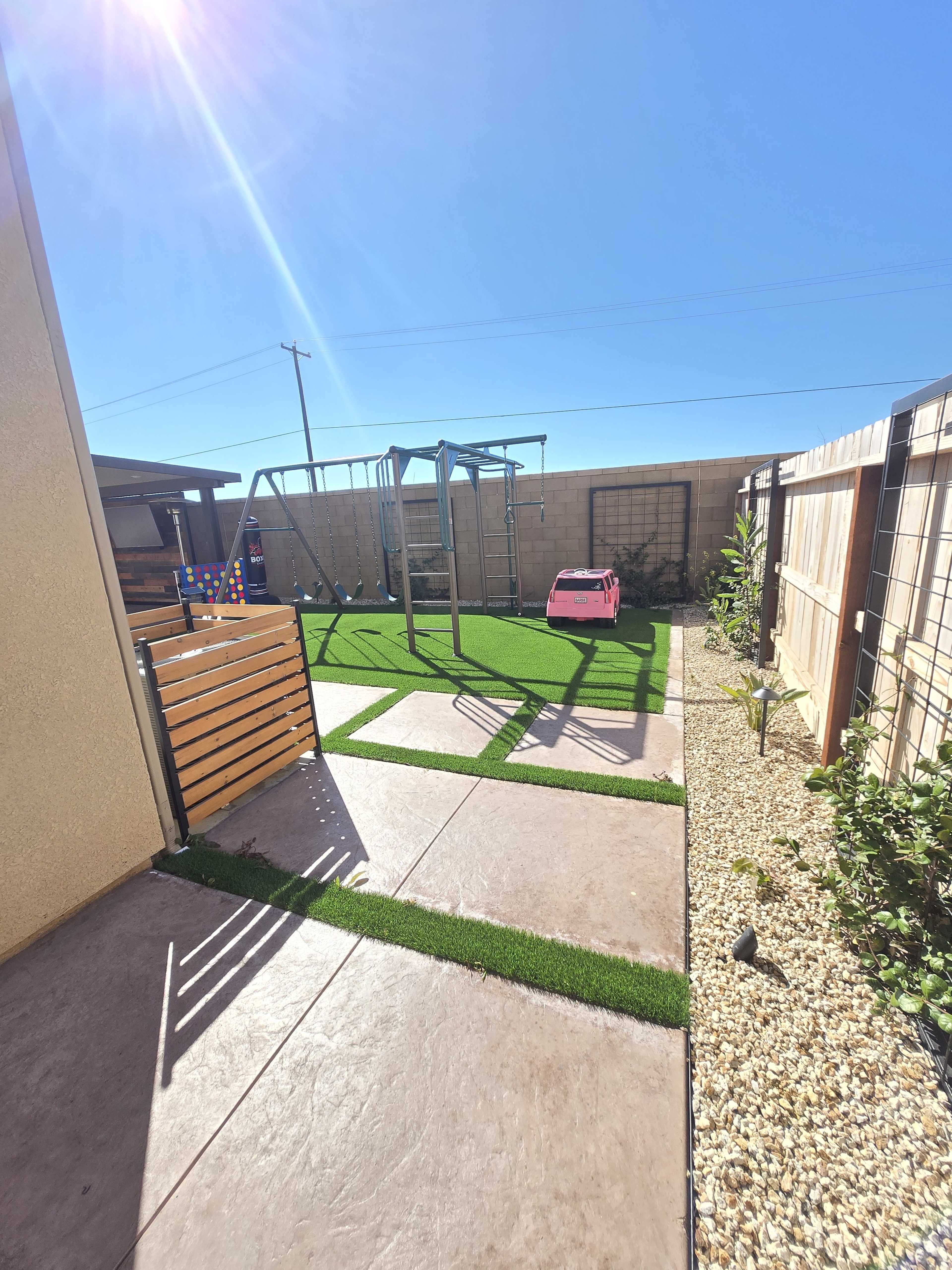 The image depicts a backyard with artificial grass, a swing set, a small pink play car, and gravel landscaping.