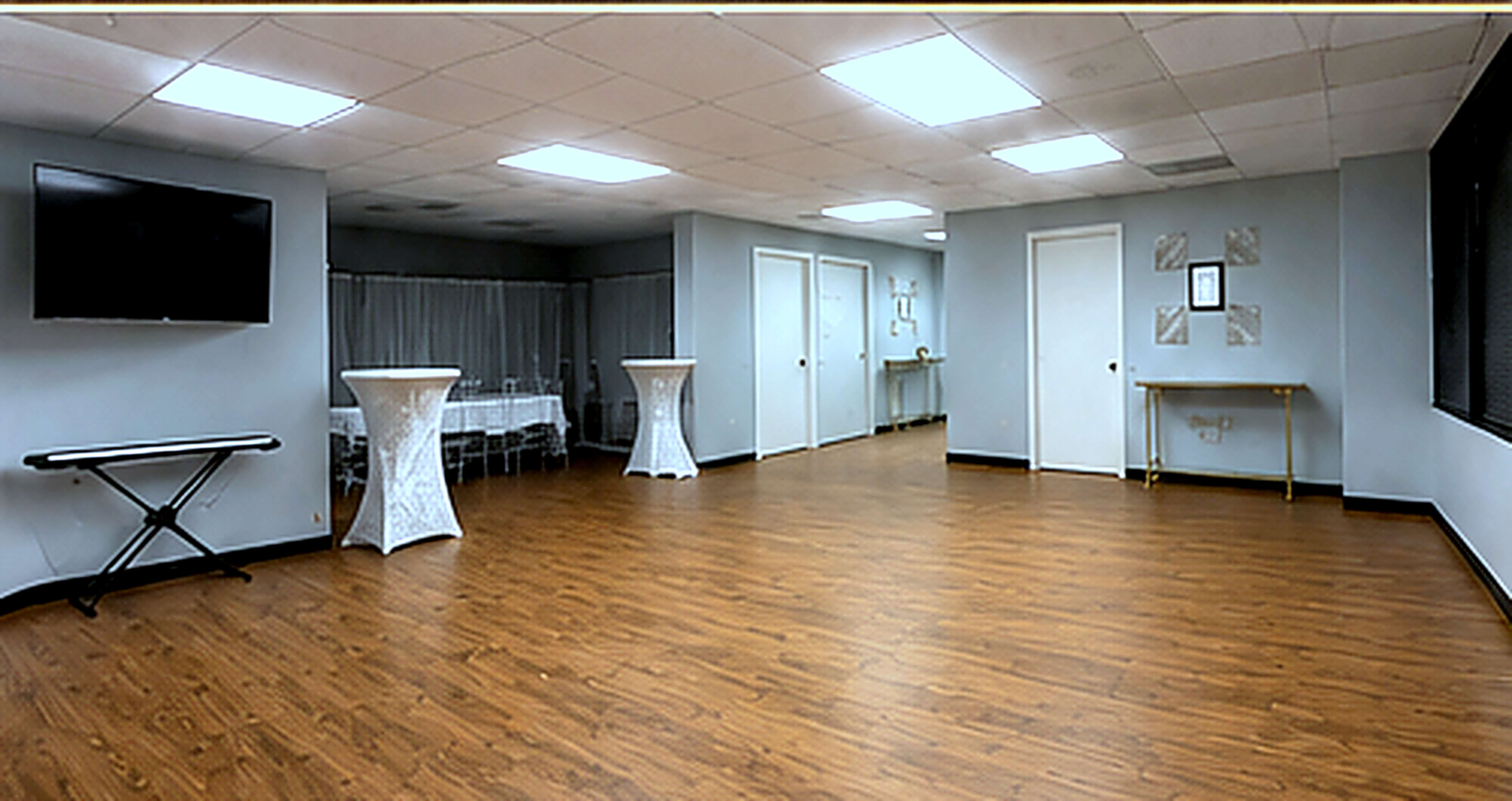 The image shows a spacious, well-lit event room with wooden flooring, several tables with white tablecloths in the background, and a keyboard in the corner.