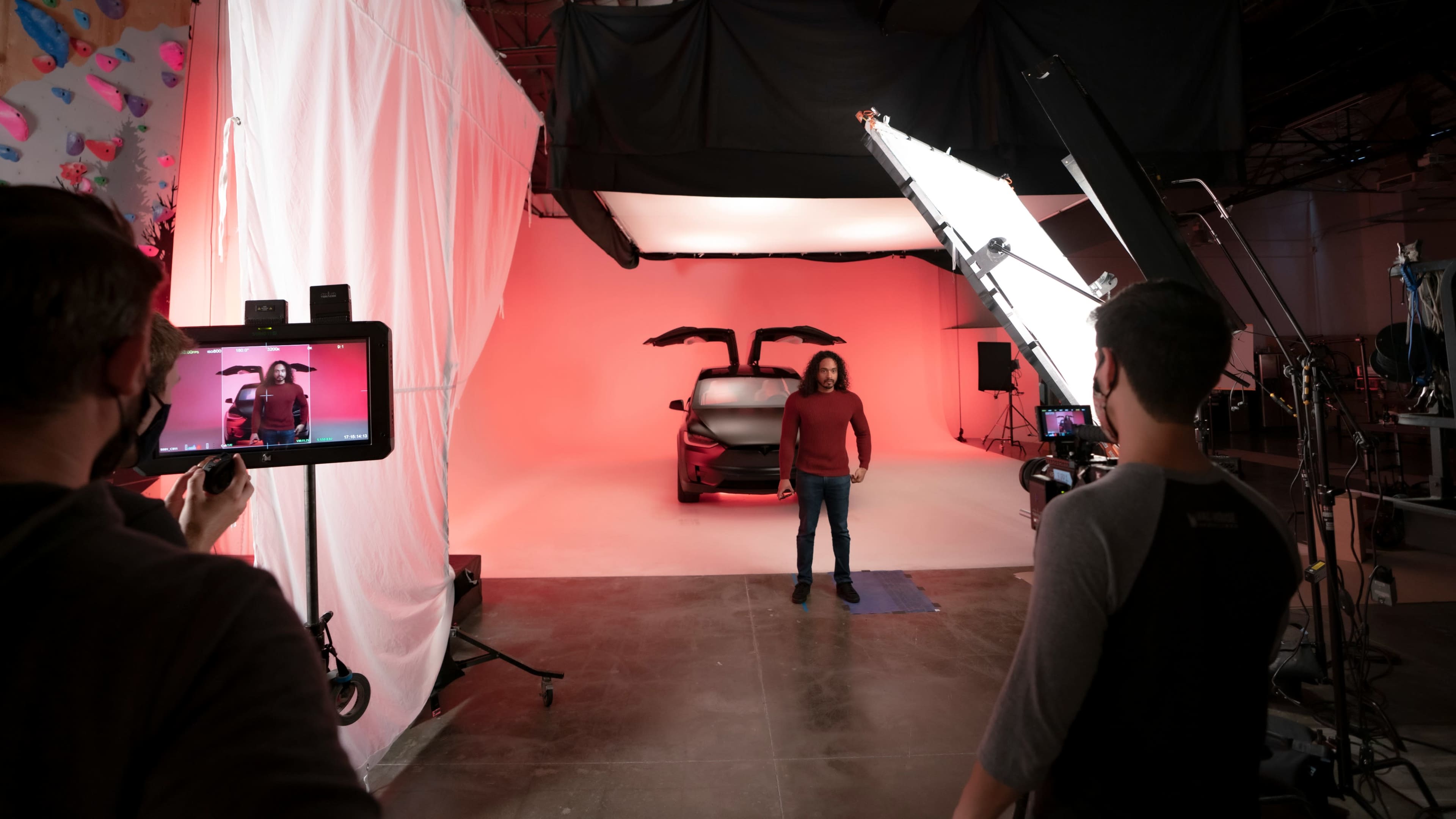 A person stands in front of a car with falcon-wing doors in a studio setting, while two crew members operate cameras and lighting equipment.