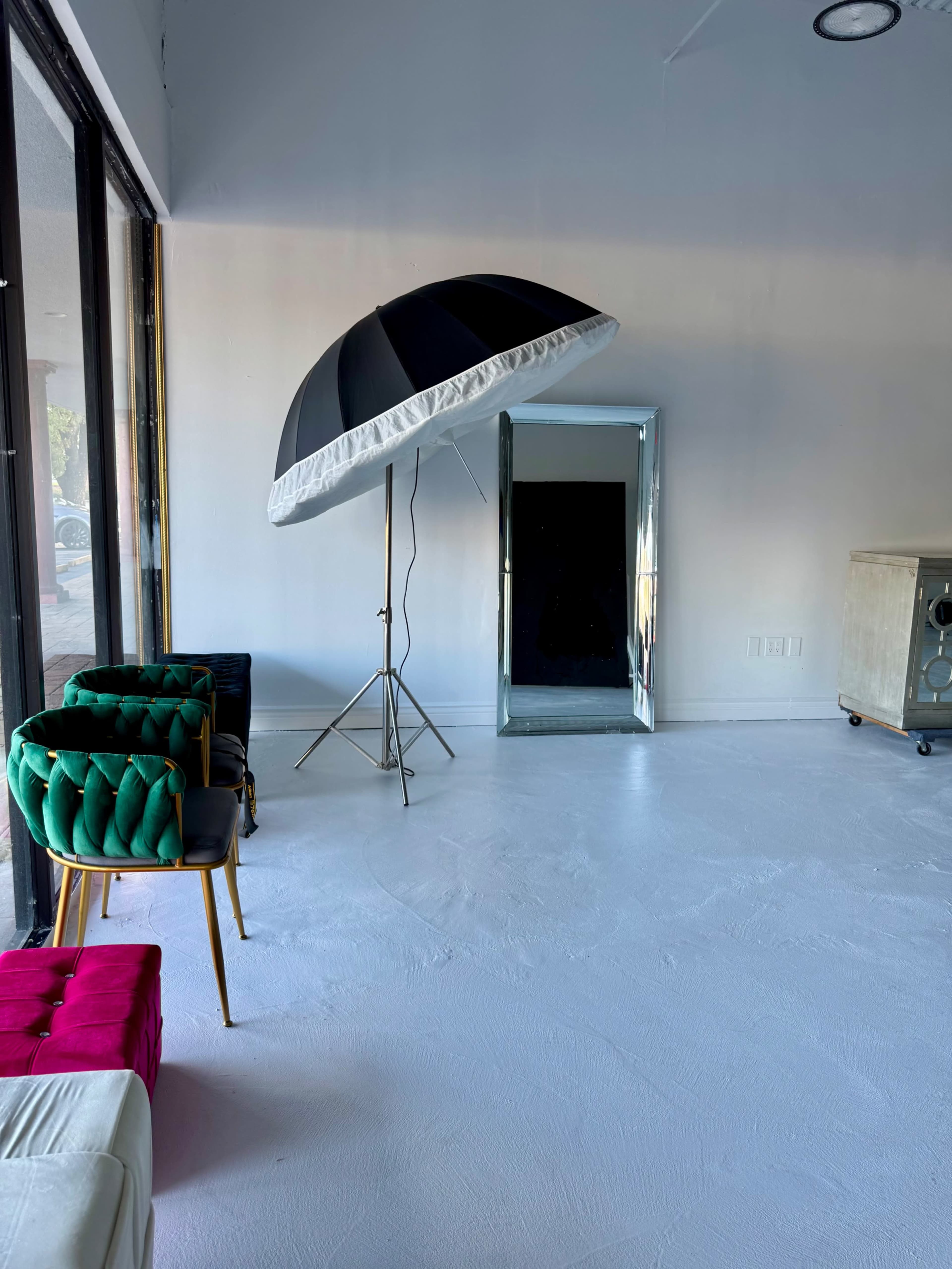The image shows a minimalistic interior space with a large black and white umbrella light stand, a full-length mirror, and a few colorful chairs.