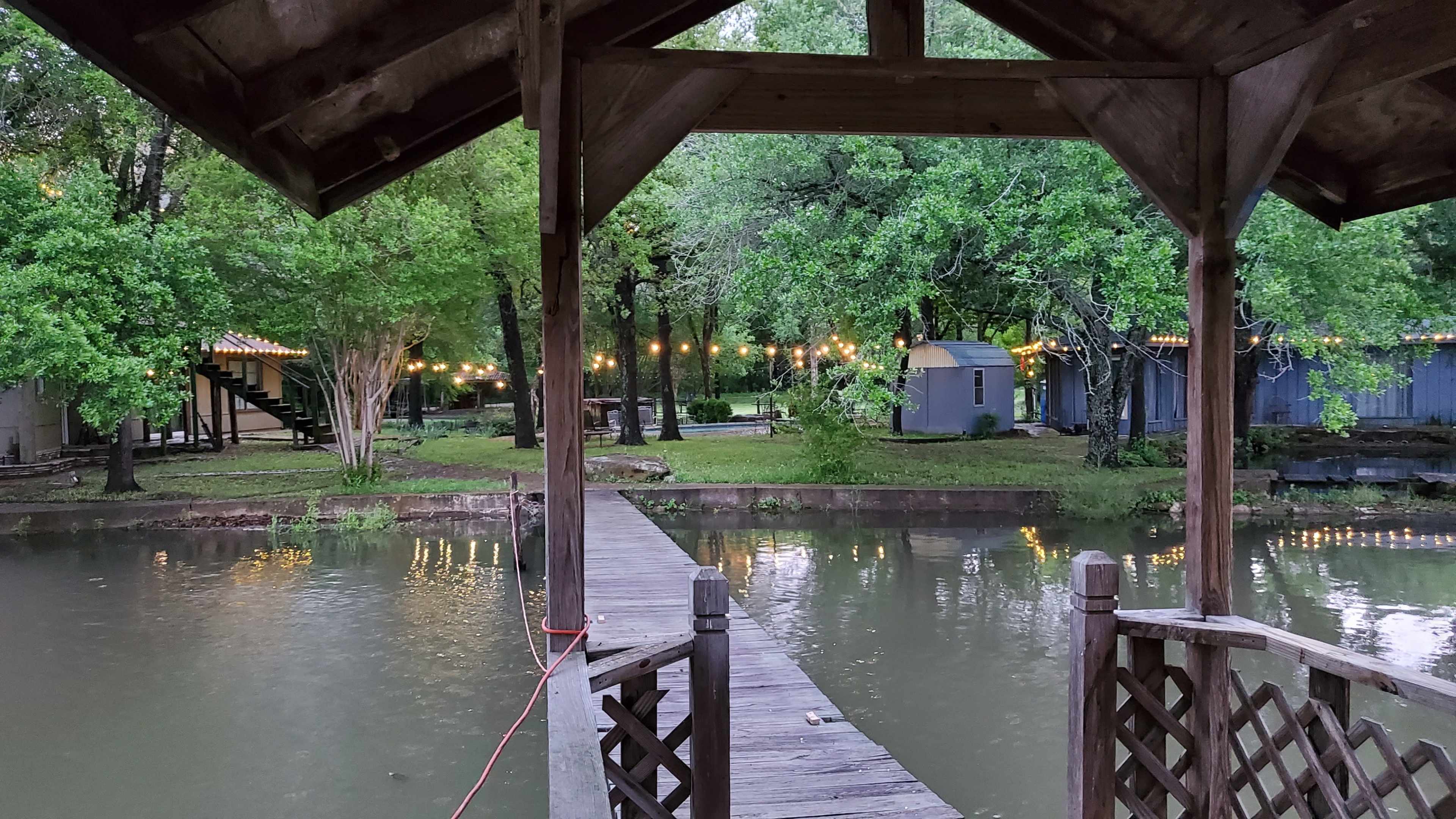 A wooden dock extends over a calm body of water, surrounded by trees and cabins, with decorative lights illuminating the scene.
