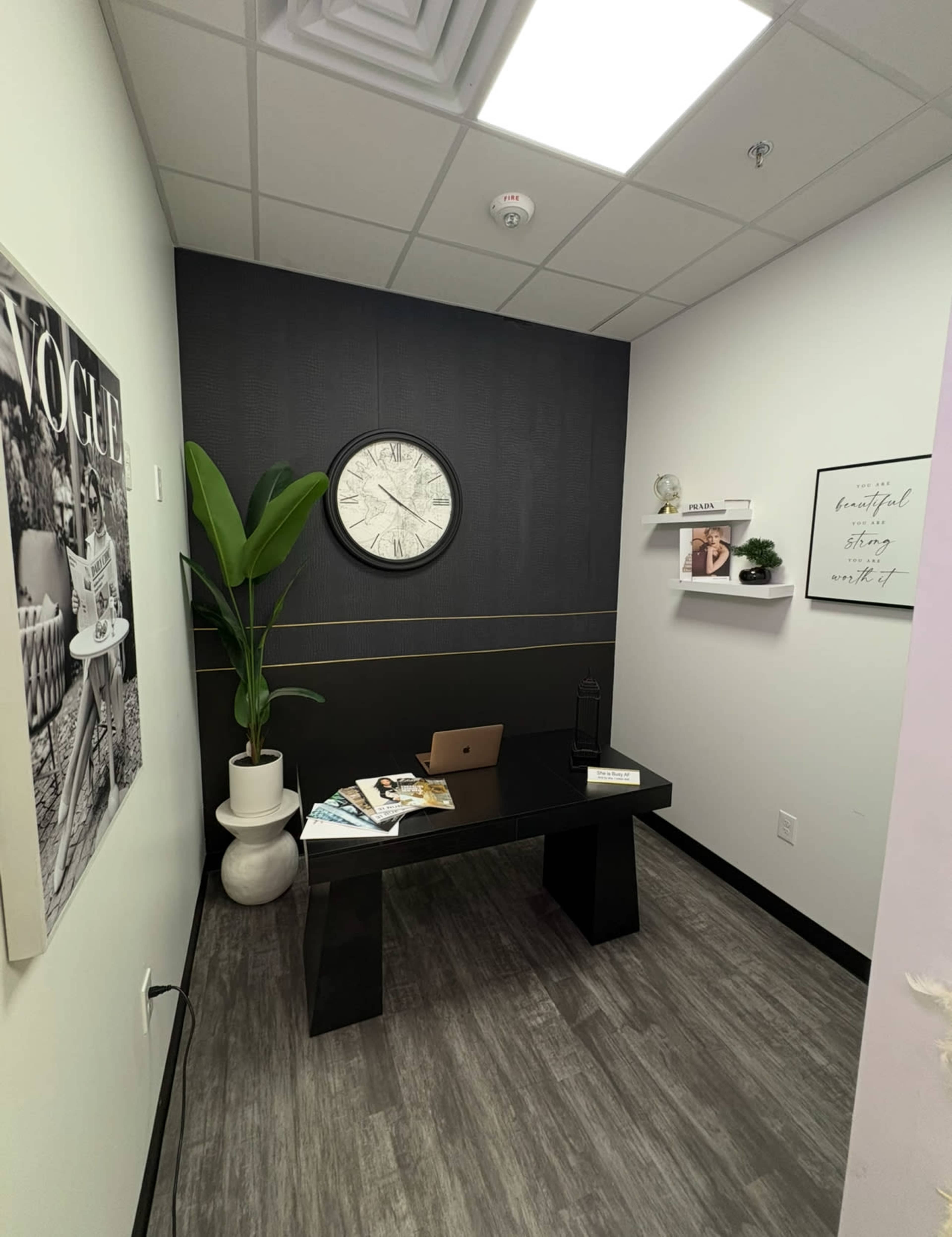 The image shows a small office space featuring a black desk, a clock, decorative shelves, a potted plant, and various magazines on the desk.