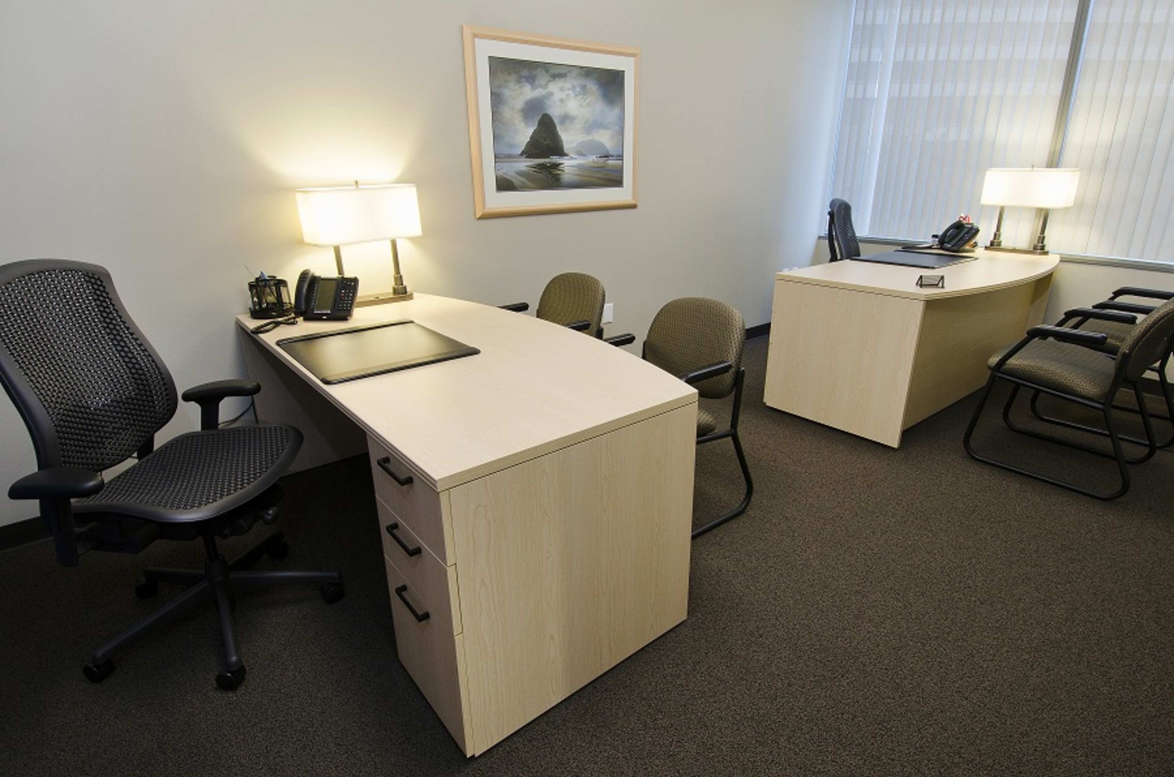 The image shows a modern office space with two desks, an office chair, and a waiting area with stackable chairs, all illuminated by lamps and natural light.