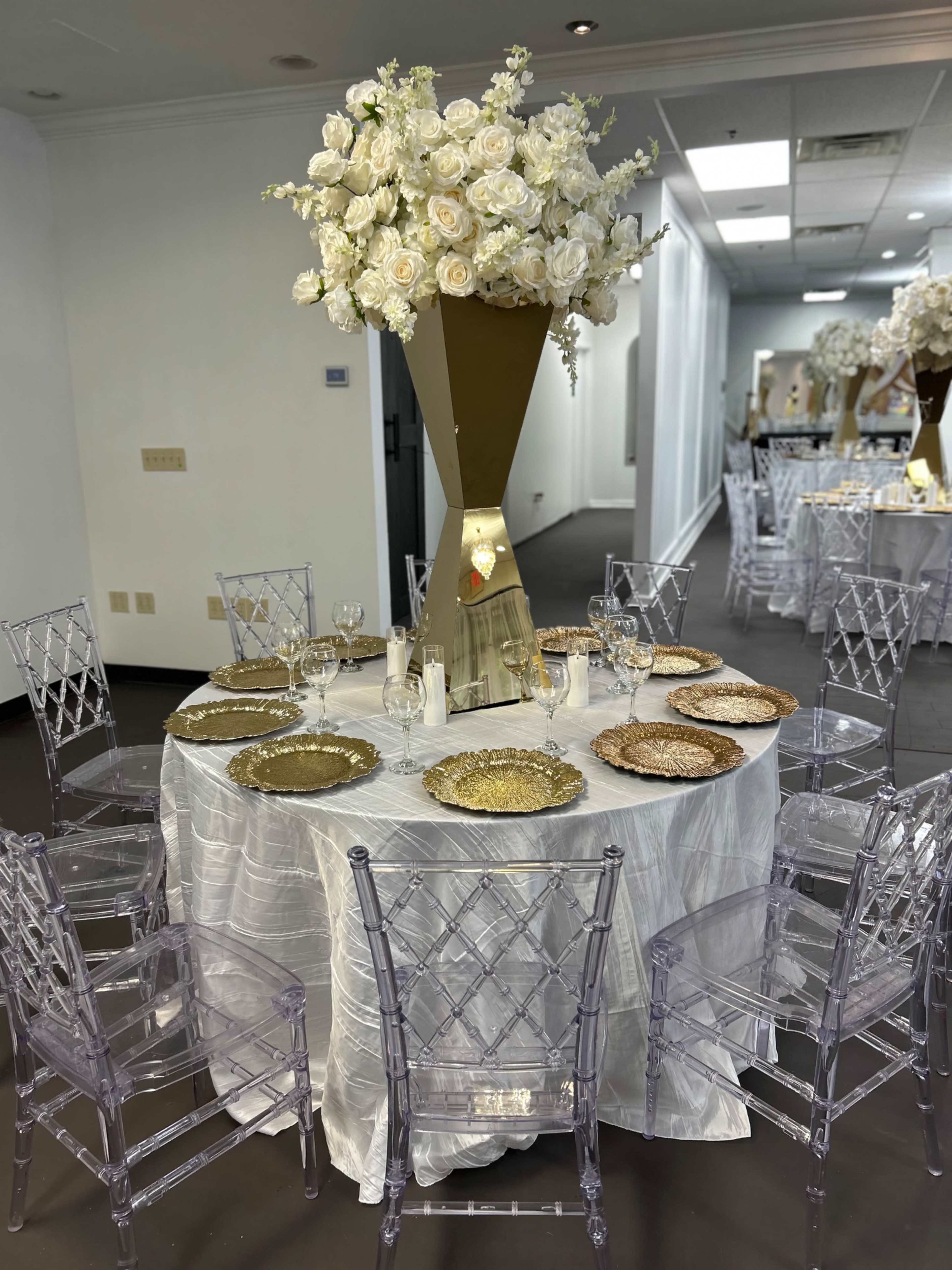A round dining table is set with gold chargers and crystal glassware, surrounded by transparent acrylic chairs, and adorned with a tall floral arrangement of white roses in a gold vase.