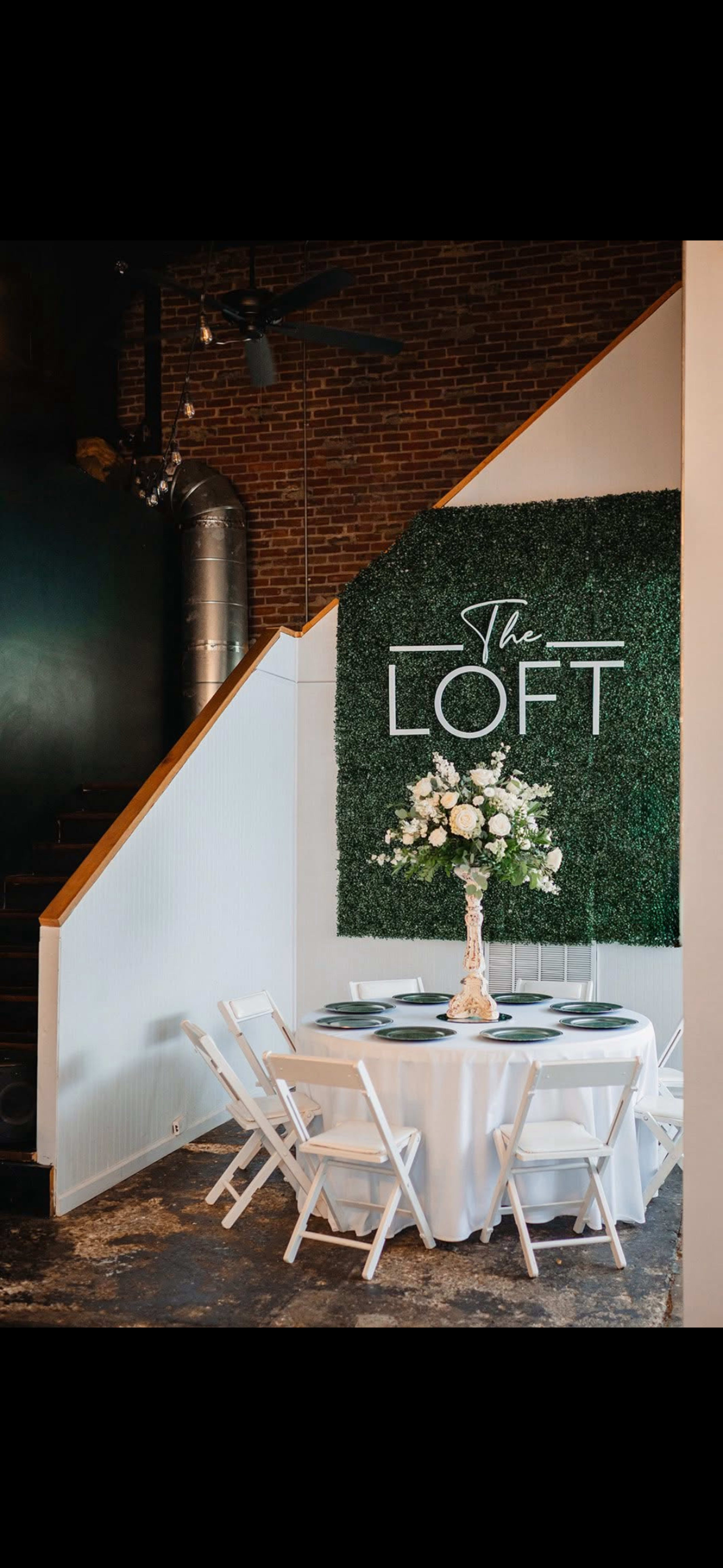 A round table with a white tablecloth and floral centerpiece is set up in front of a green wall that displays the name "The Loft."