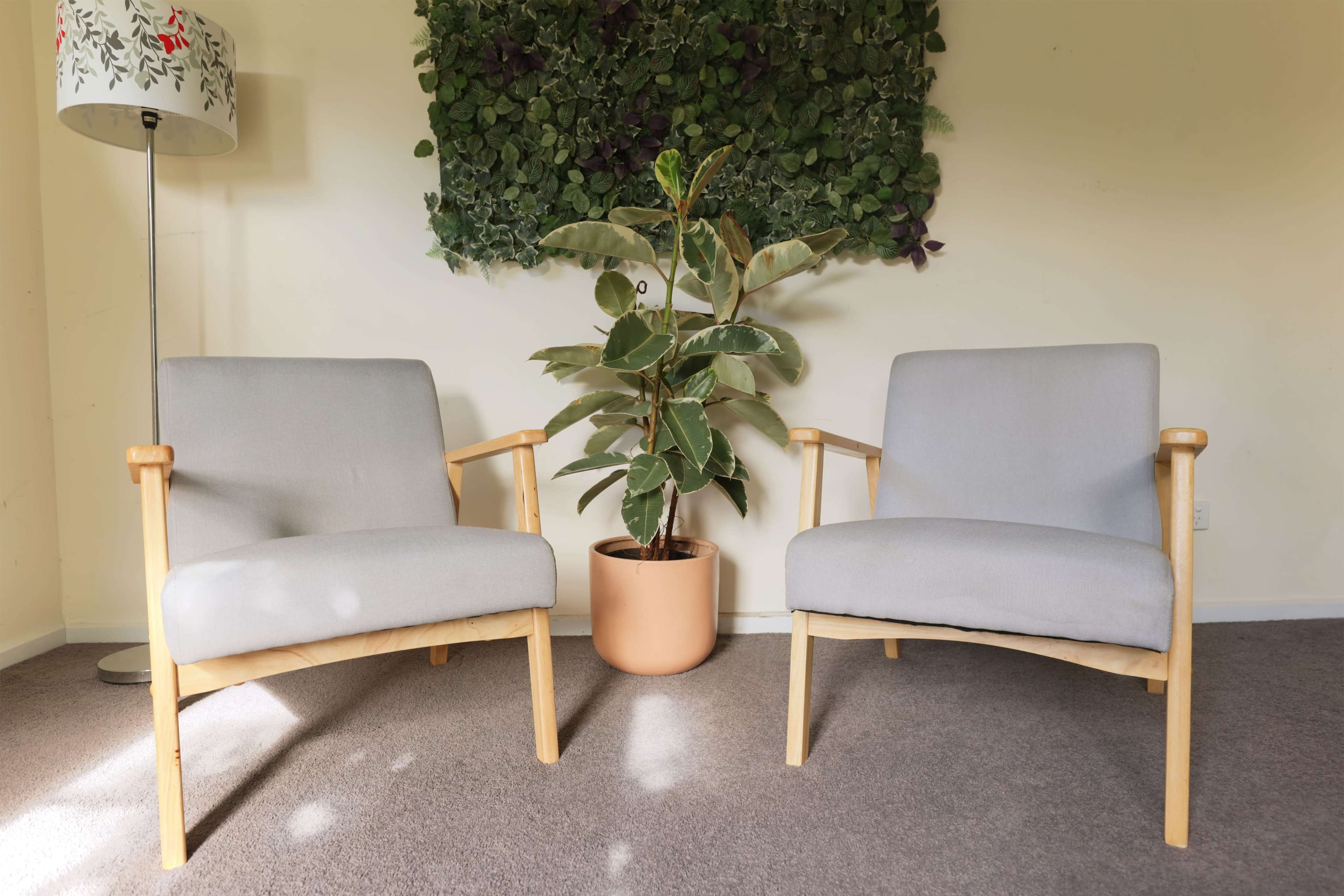 Two light gray chairs are positioned facing a potted plant against a green wall decoration in a room with carpeted flooring.