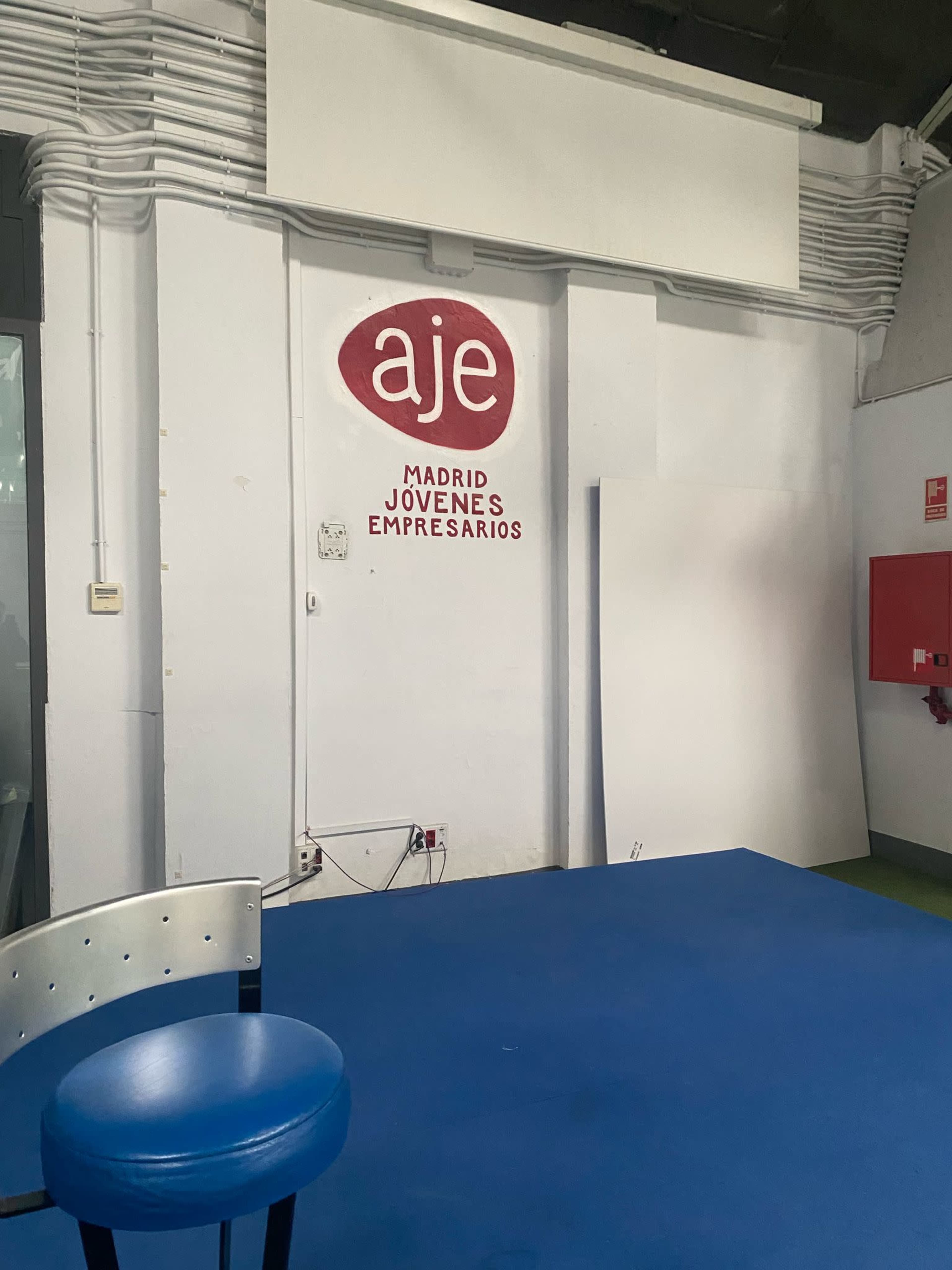 The image shows a blue table and a blue chair against a white wall with a red logo that reads "aje Madrid Jóvenes Empresarios."