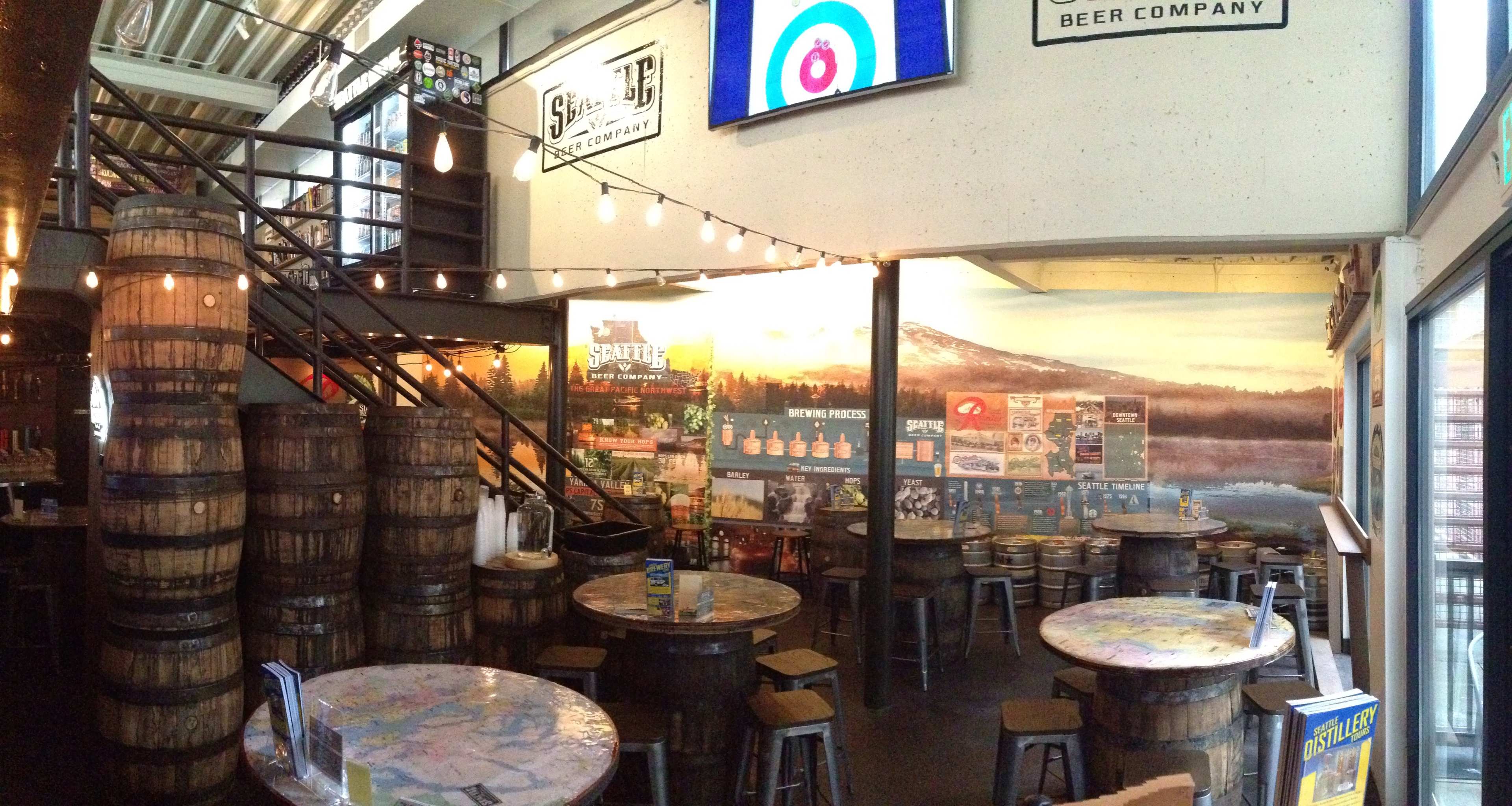 A brewery interior with wooden barrels, tables, and a staircase, featuring a mural of a mountain landscape on the back wall.