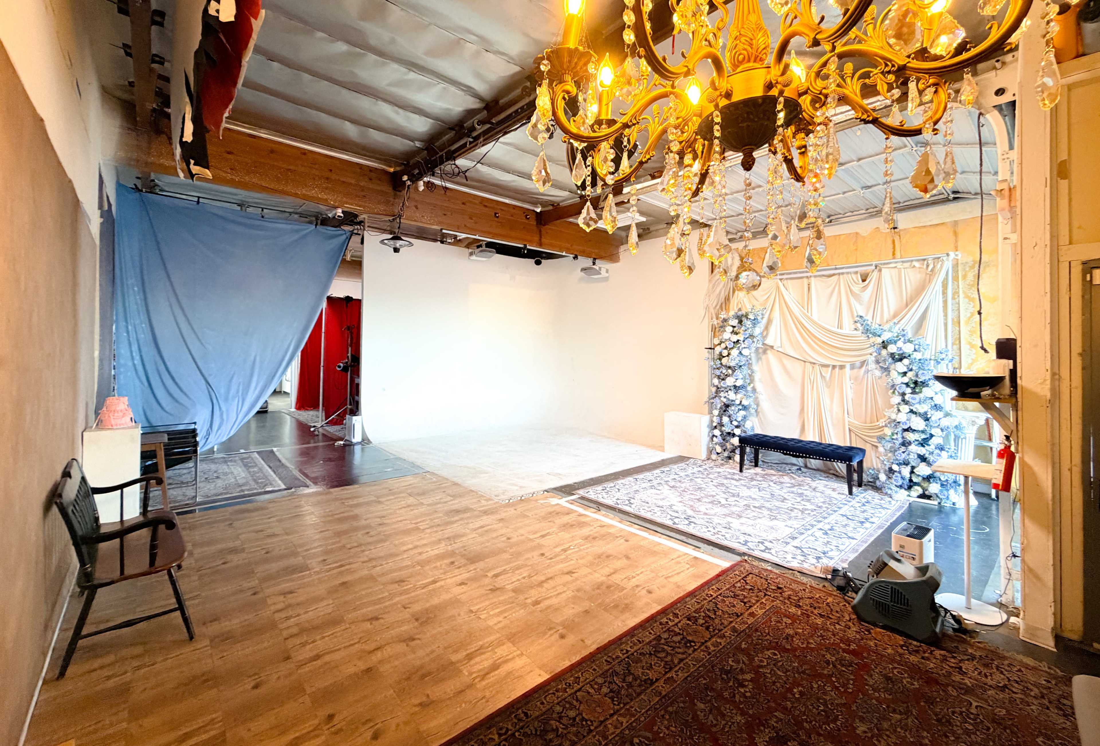 The image shows a spacious studio with a wood floor, featuring a blue backdrop on one side, a red curtain on another, and decorative elements like a chandelier and Christmas trees with white decorations.