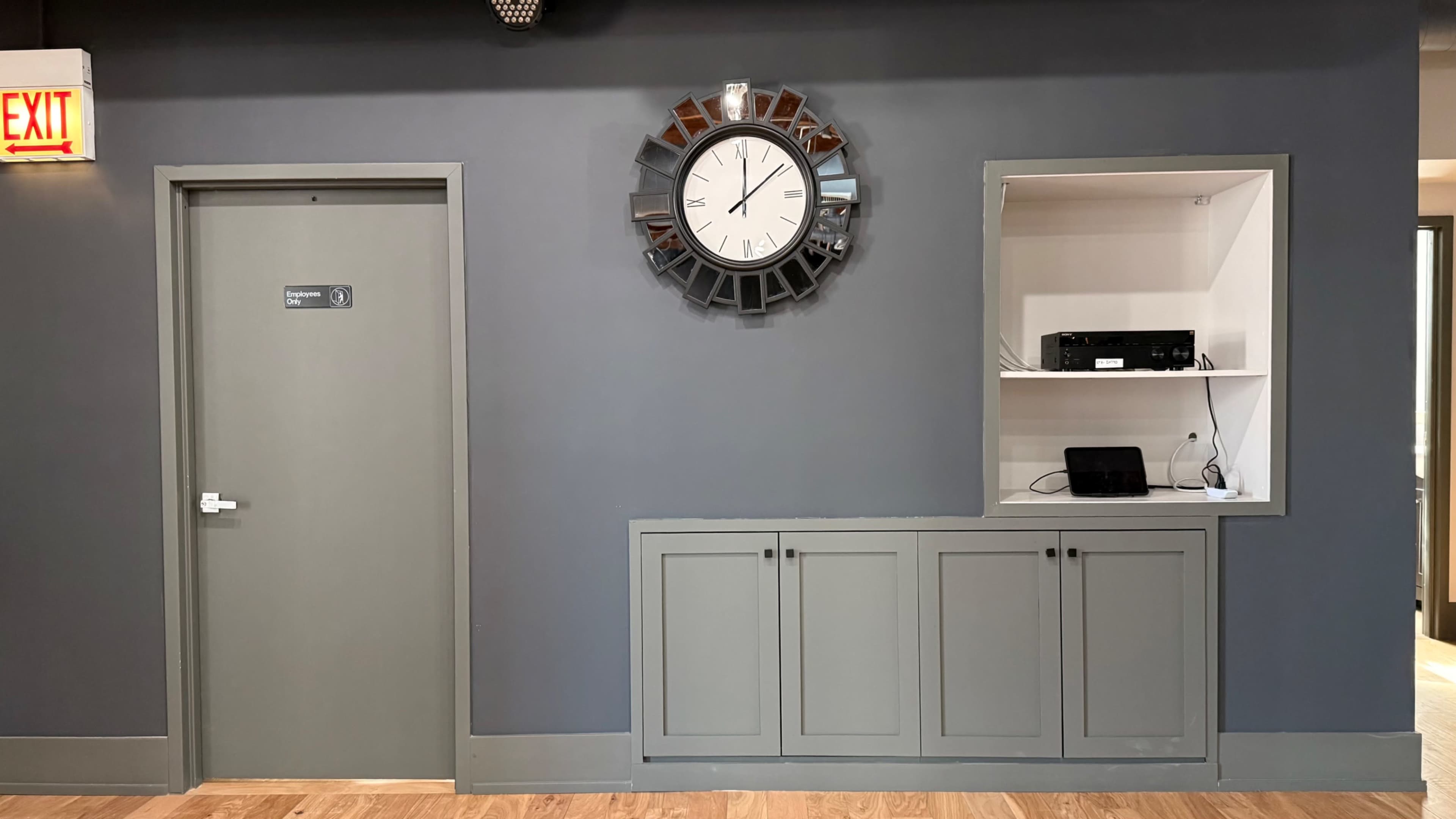 The image shows a gray wall featuring a large clock above a shelf with electronic equipment, next to a door marked with a sign.