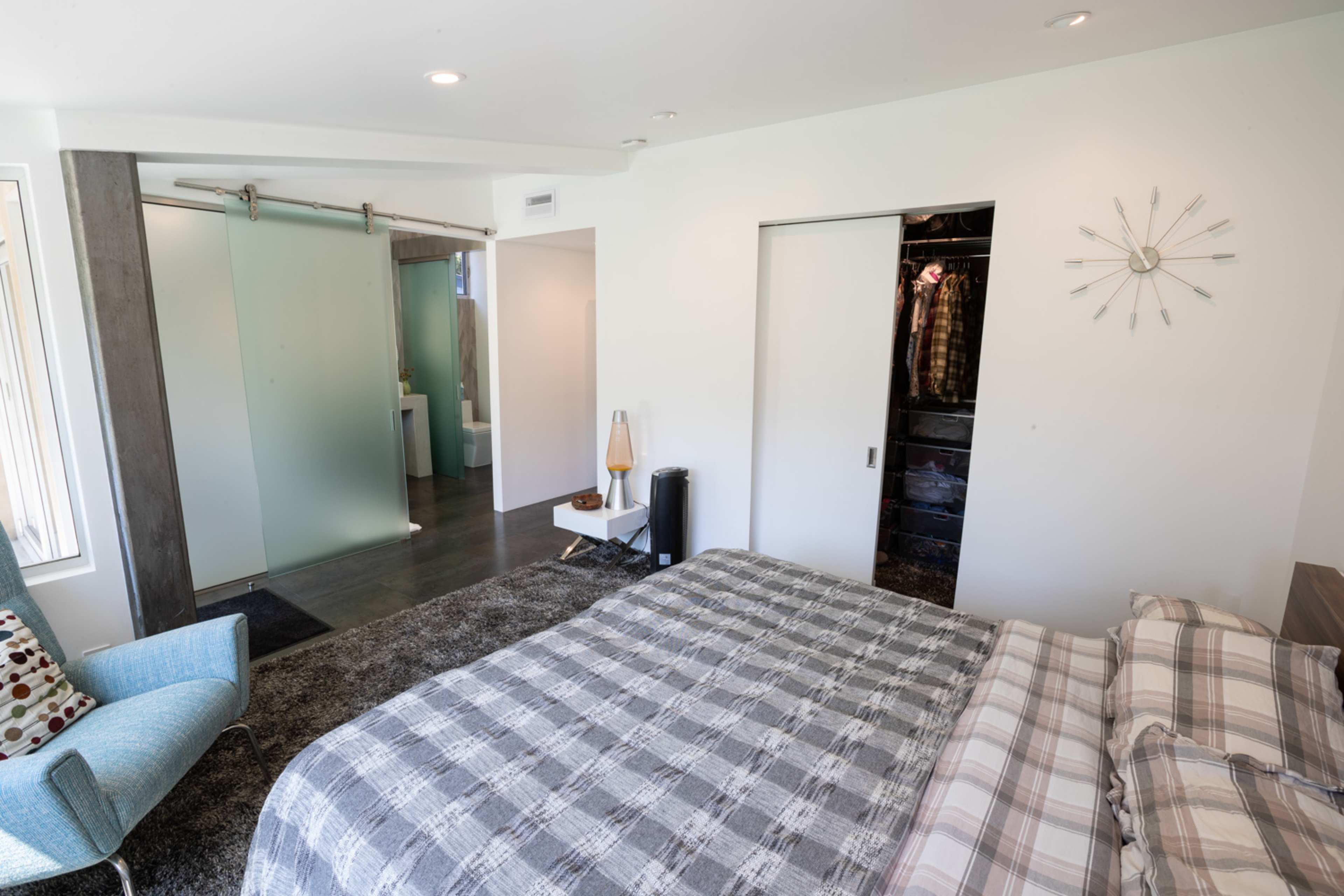 A modern bedroom features a neatly made bed with a plaid duvet, a sliding glass door leading to a bathroom area, and a chair beside a small side table.