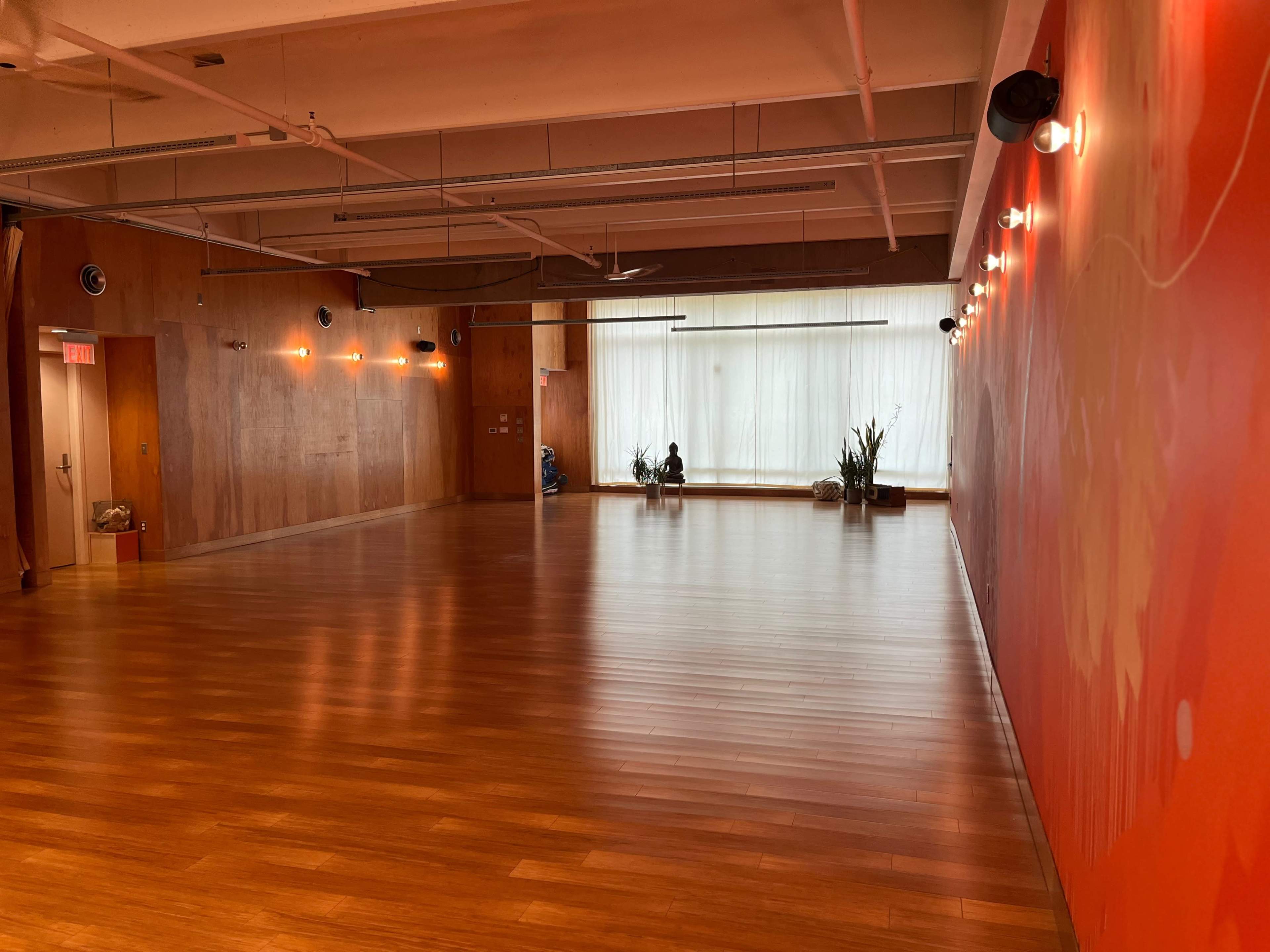 A spacious wooden-floored room with large windows and potted plants along the wall.