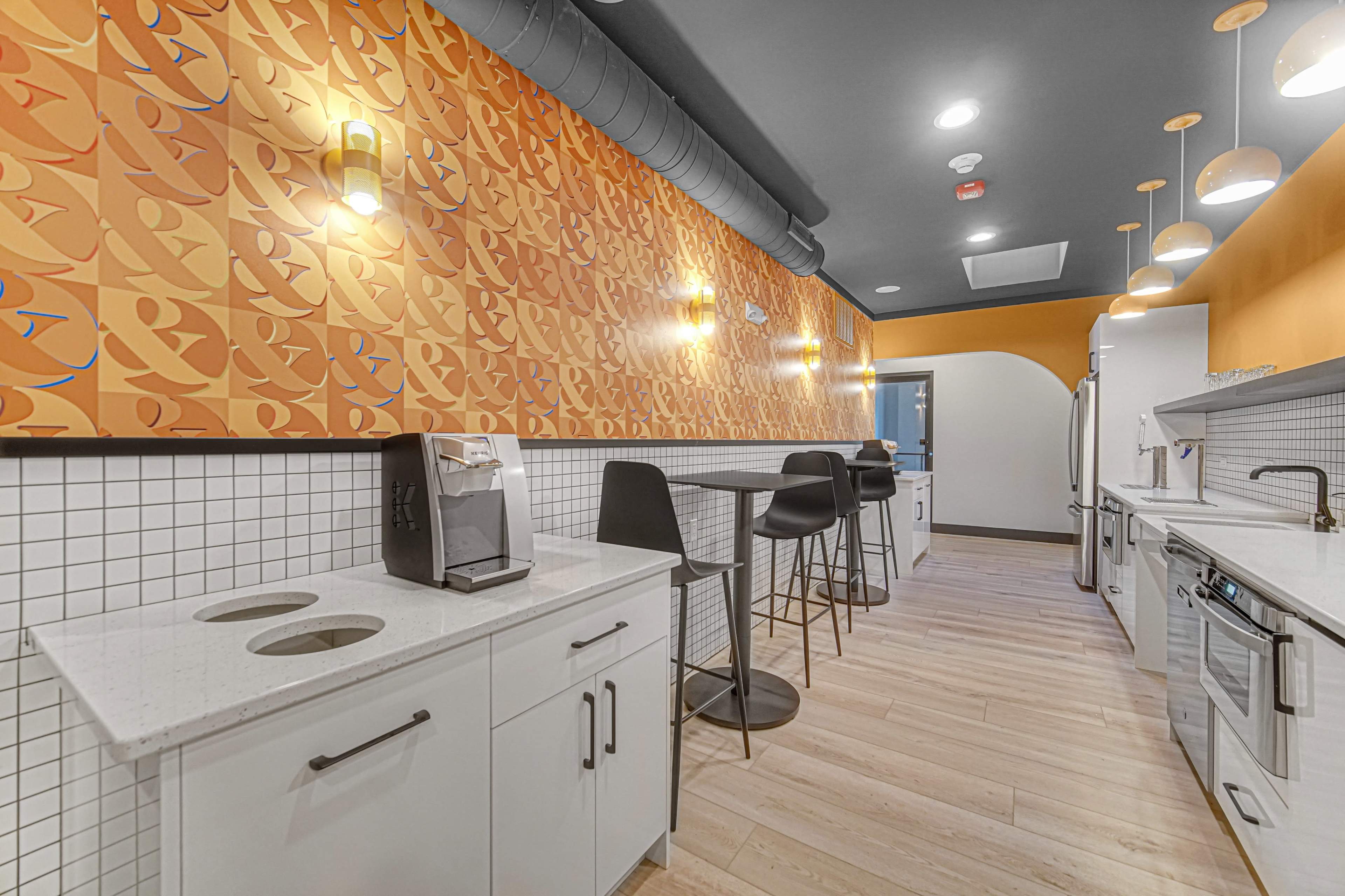 The image shows a modern kitchenette with a patterned orange wall, white cabinetry, a coffee machine, and bar-style seating area.