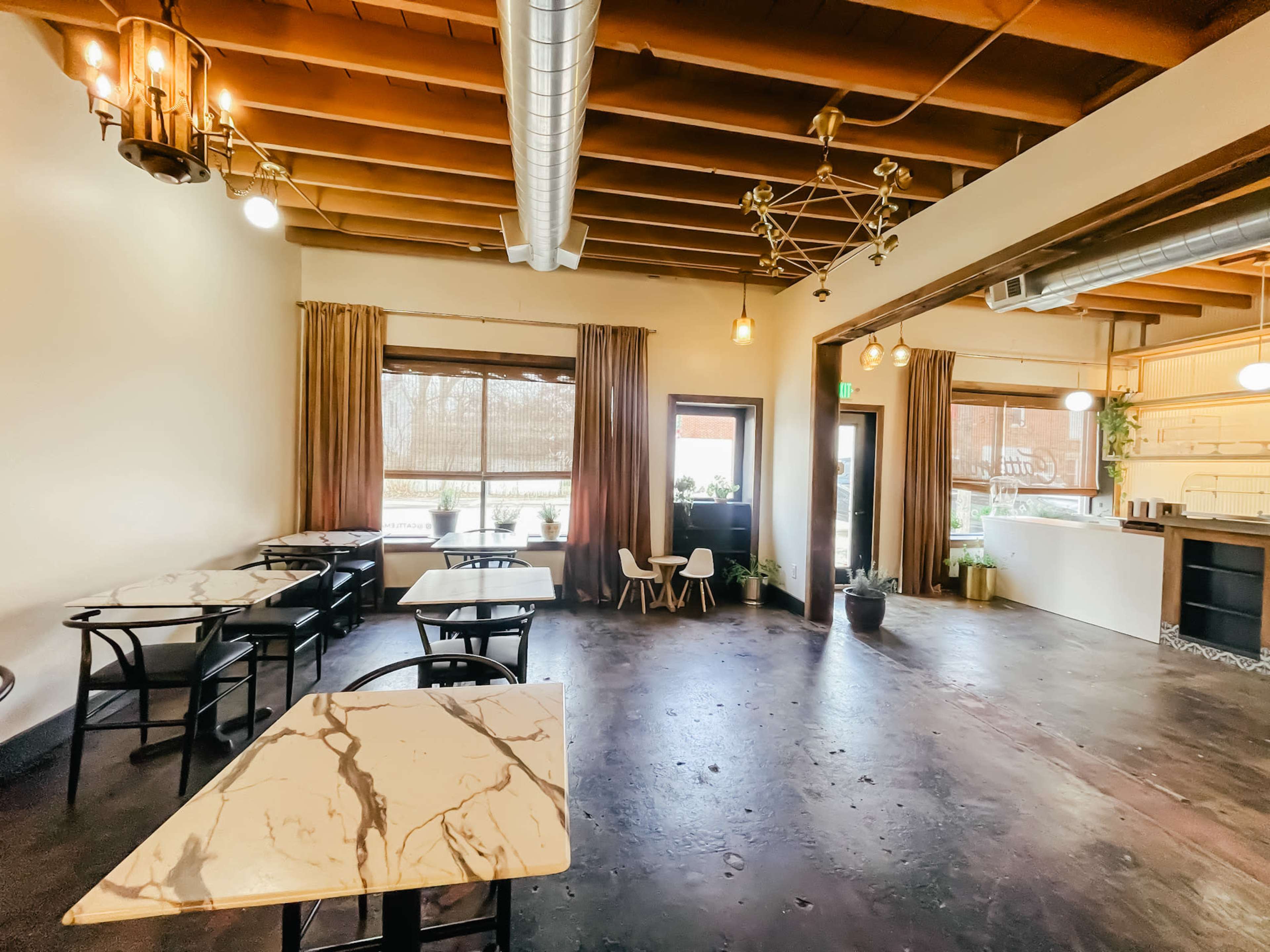 A spacious indoor area with several marble-top tables, wooden beams overhead, and large windows dressed with curtains.