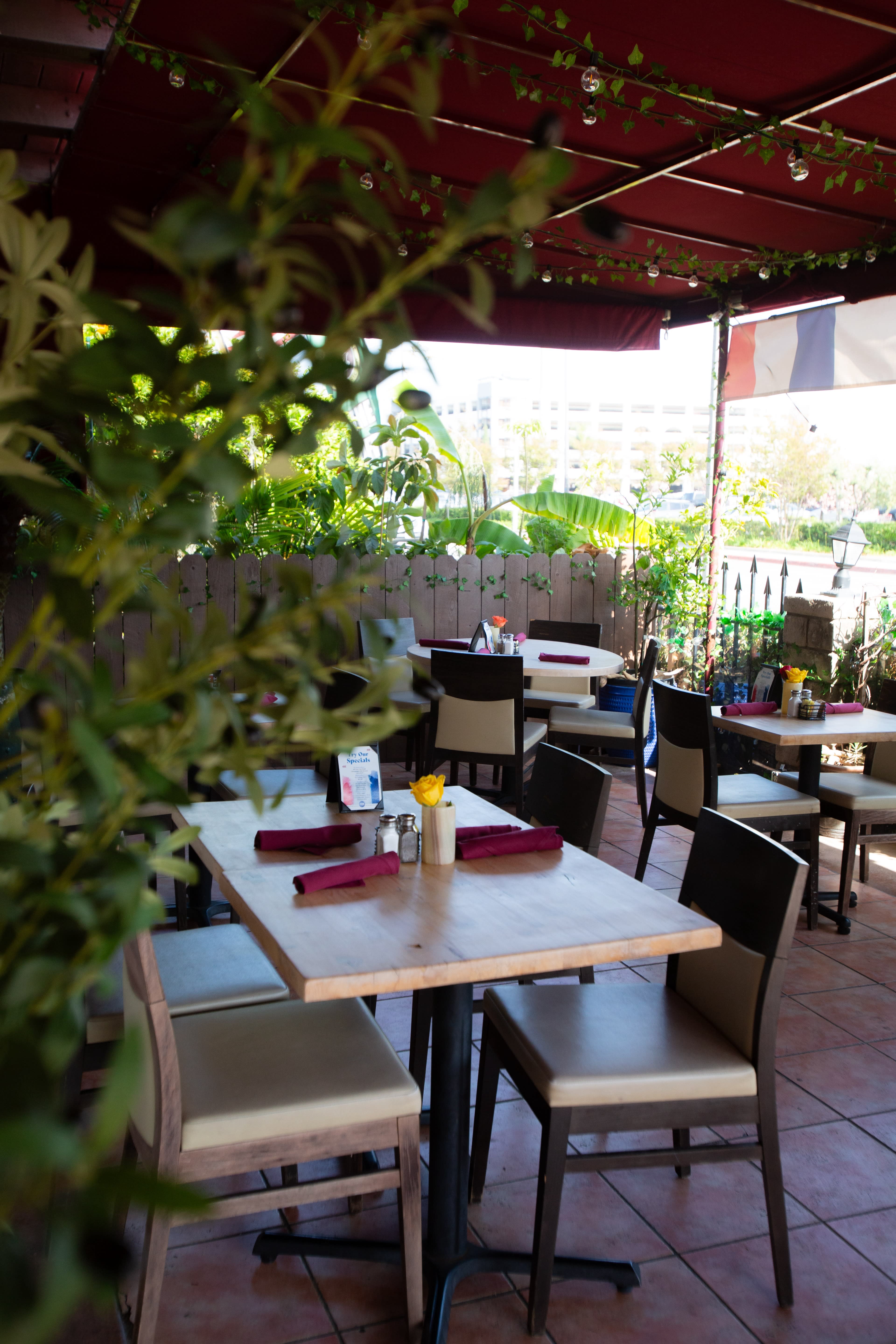 A patio dining area with tables set for guests, surrounded by plants and shaded by red awnings.