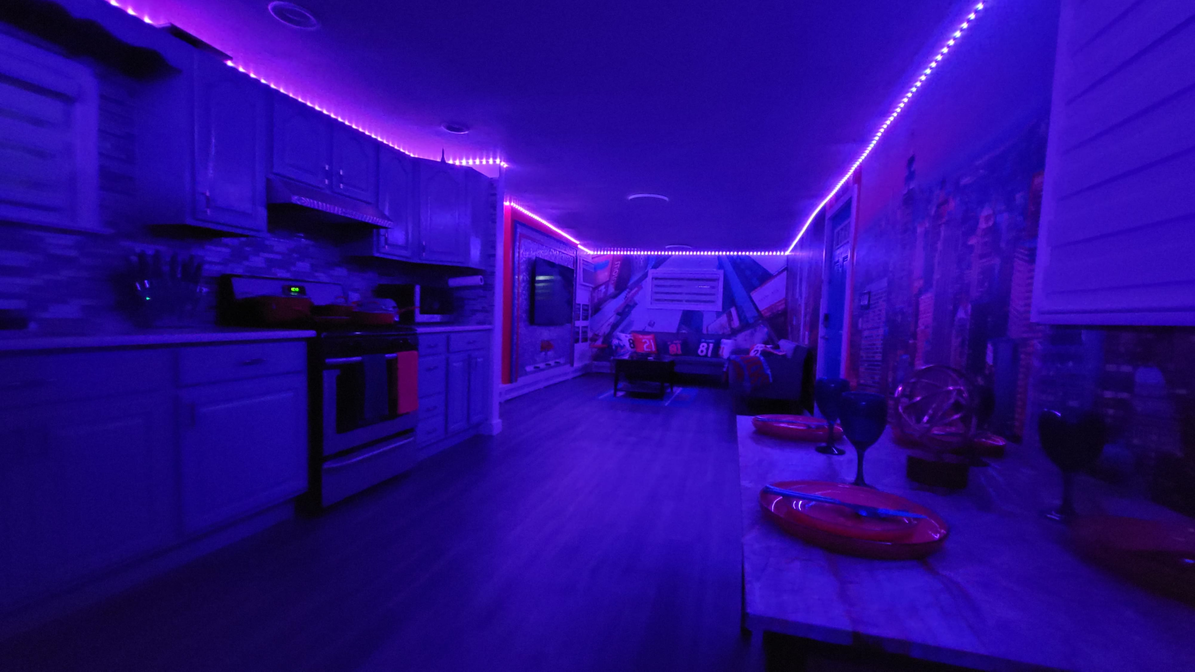 The image shows a modern kitchen and living area illuminated by purple LED lights, with contemporary furnishings and a cityscape mural on the walls.
