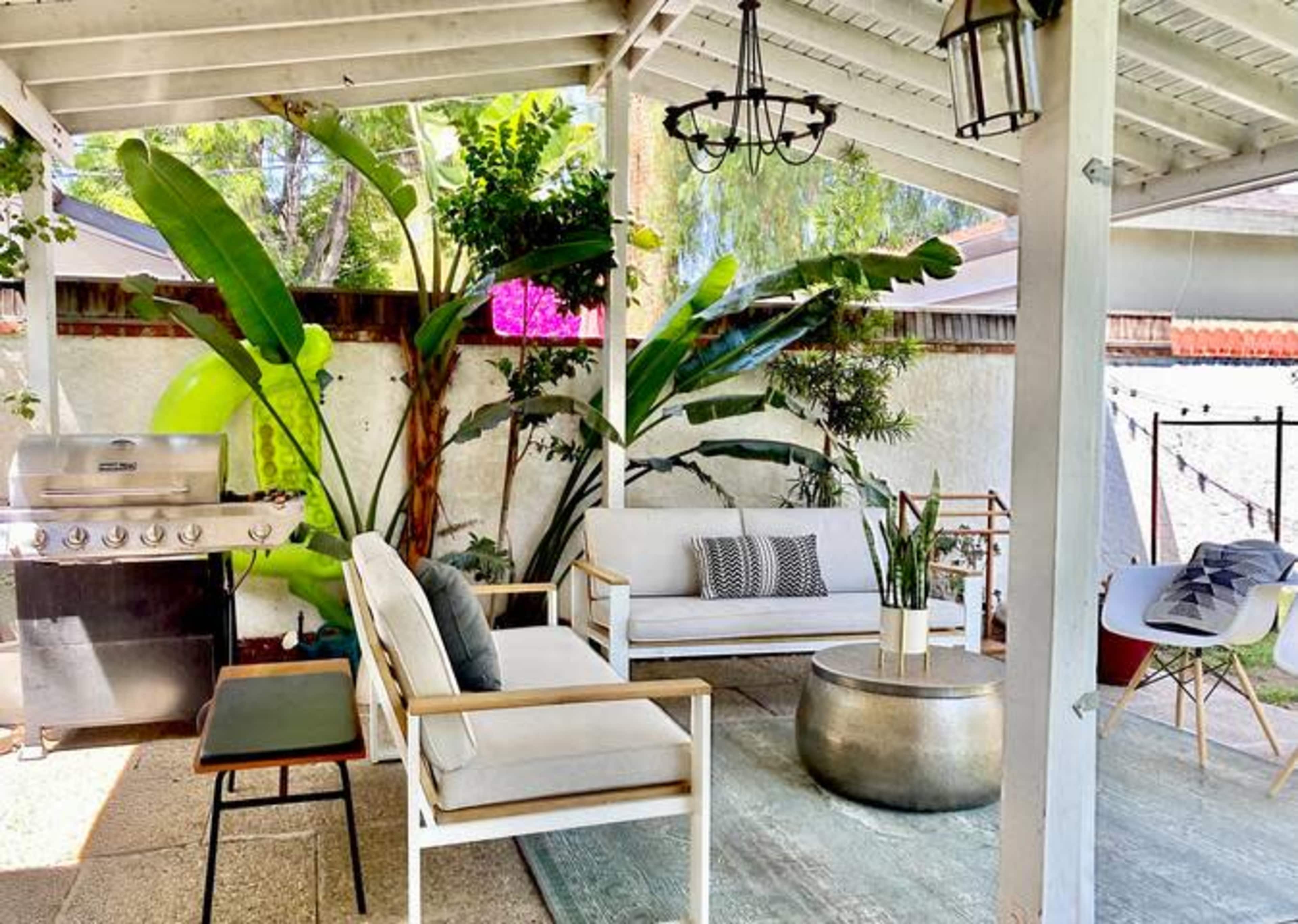 A covered outdoor seating area with a sofa, coffee table, and a grill, surrounded by tropical plants.
