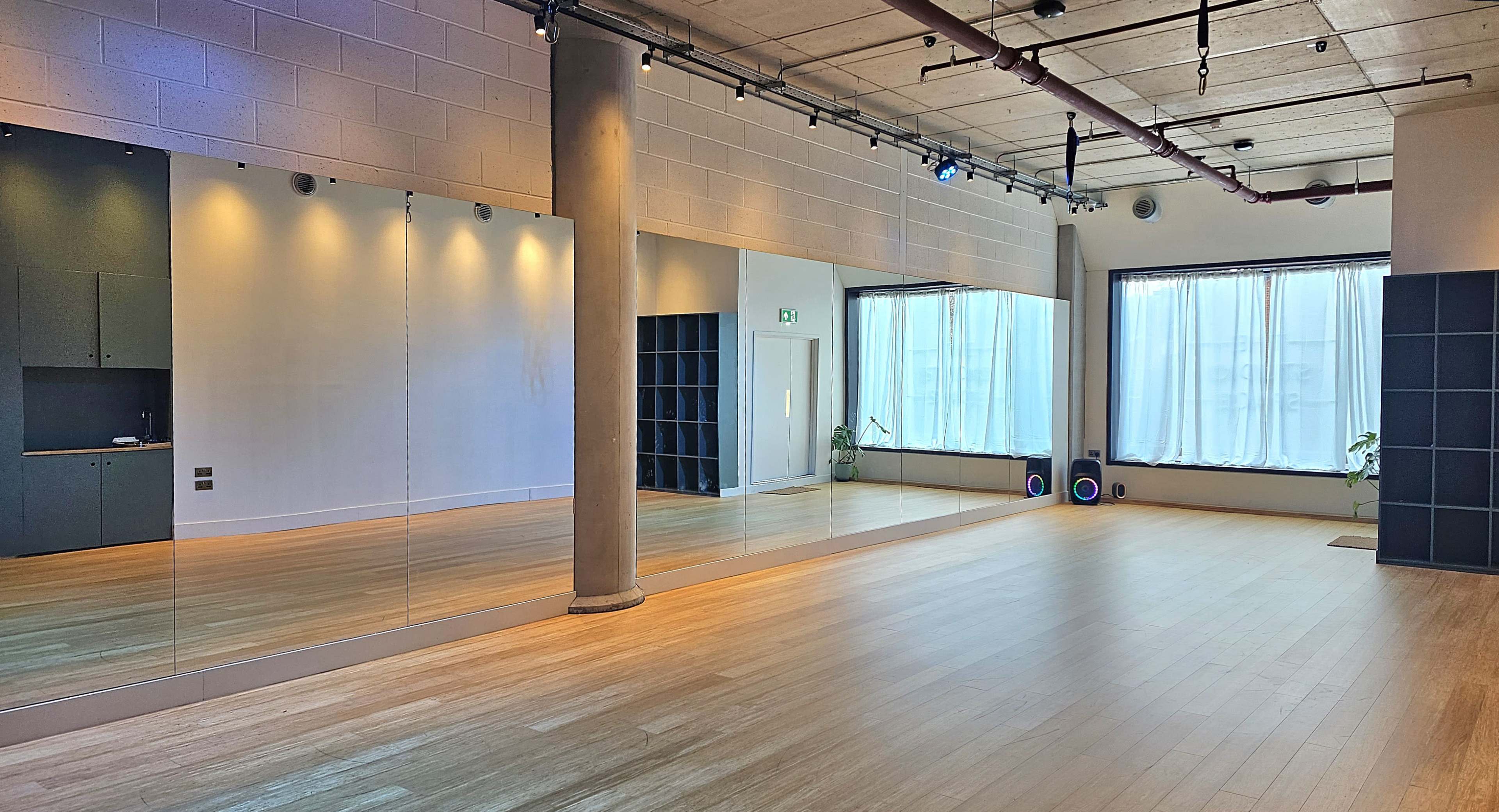 The image shows a spacious studio with large mirrors, wooden floors, and soft natural light coming through the windows.